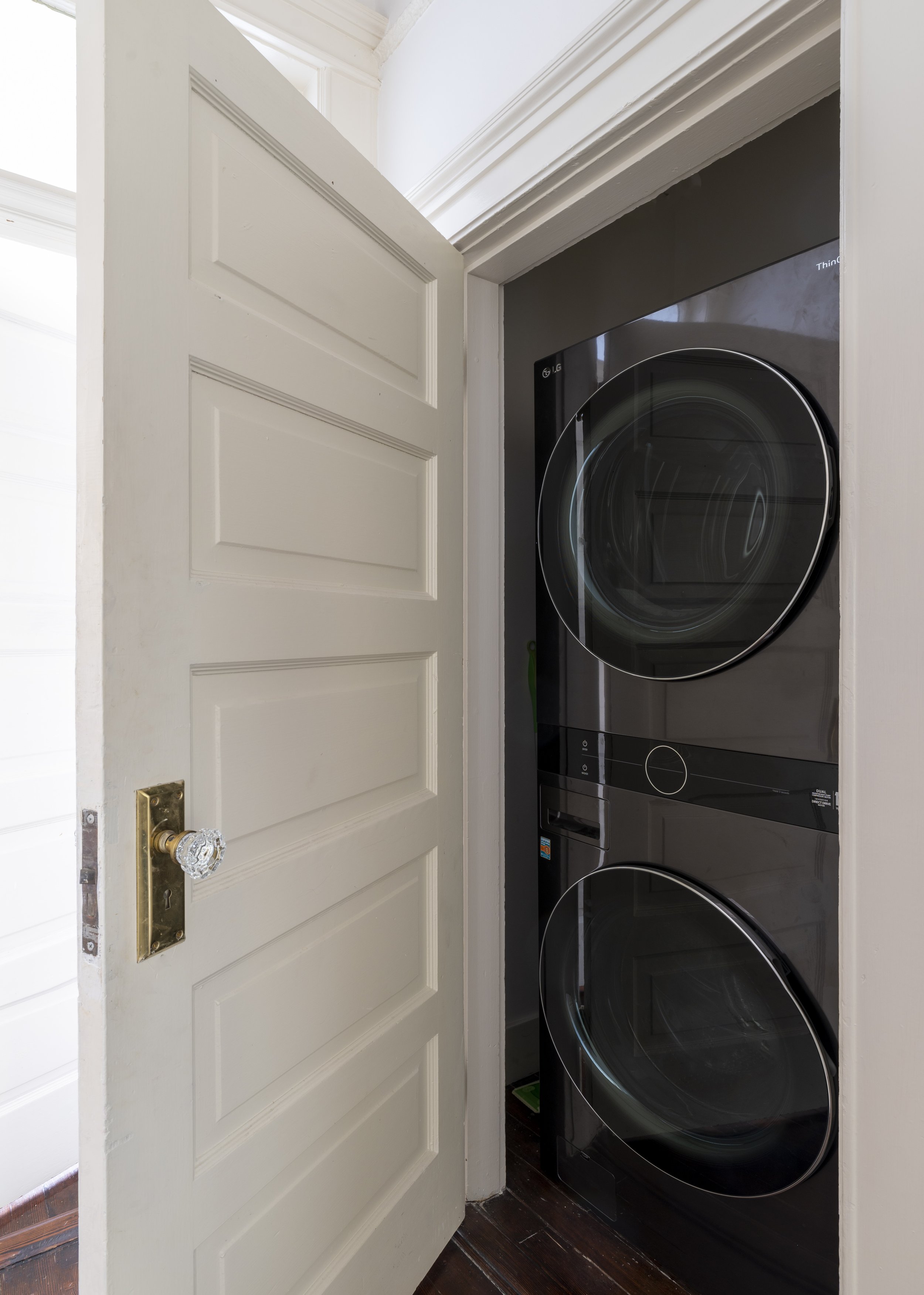 A laundry room with a white door open to reveal a black LG washer and dryer stacked vertically inside a small space. The door has a glass doorknob and a brass latch plate.