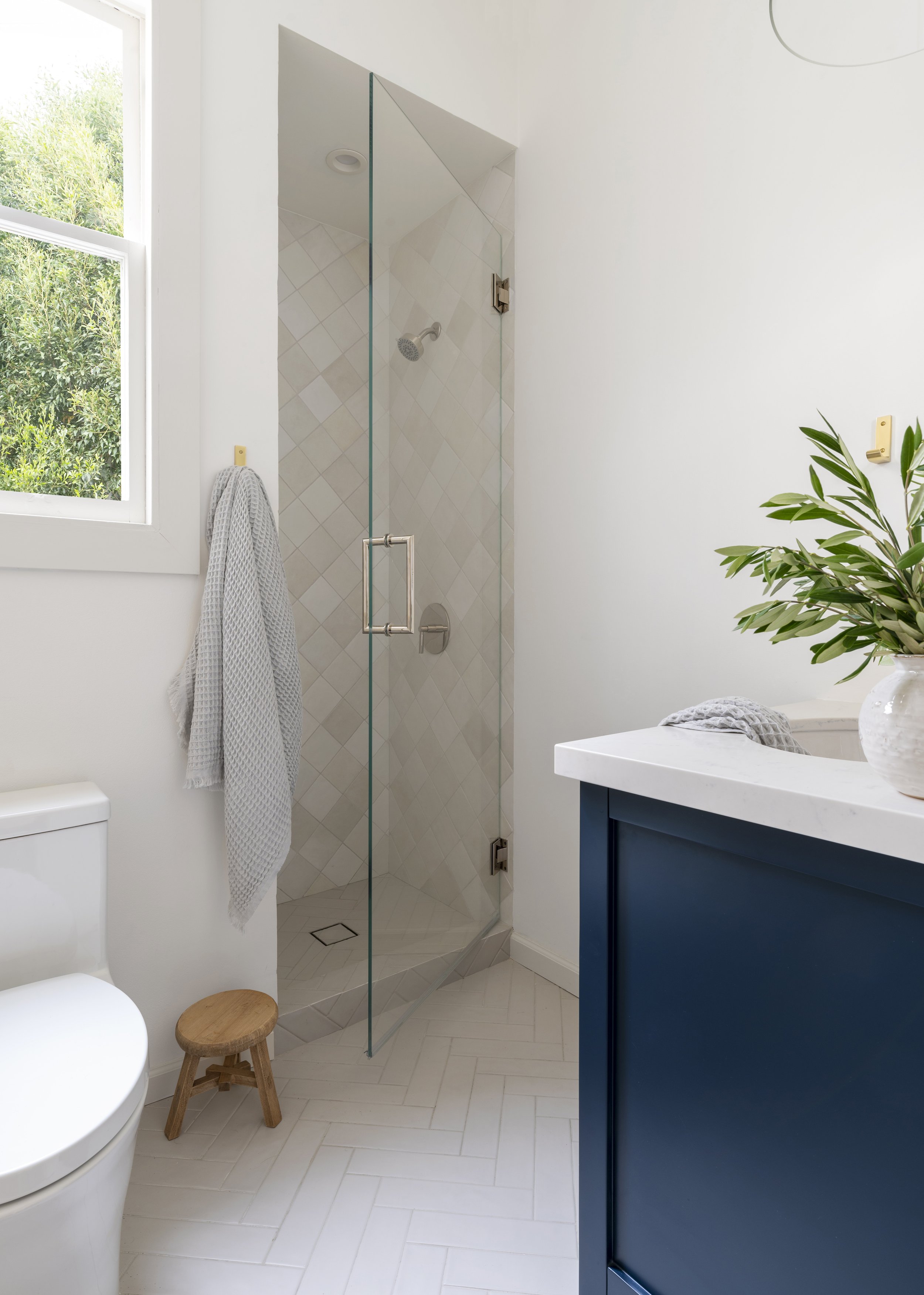 A modern bathroom with a glass shower enclosure, white walls, a window with outside greenery, a plant on the vanity, a white toilet, a small wooden stool, and a blue vanity with a white countertop.