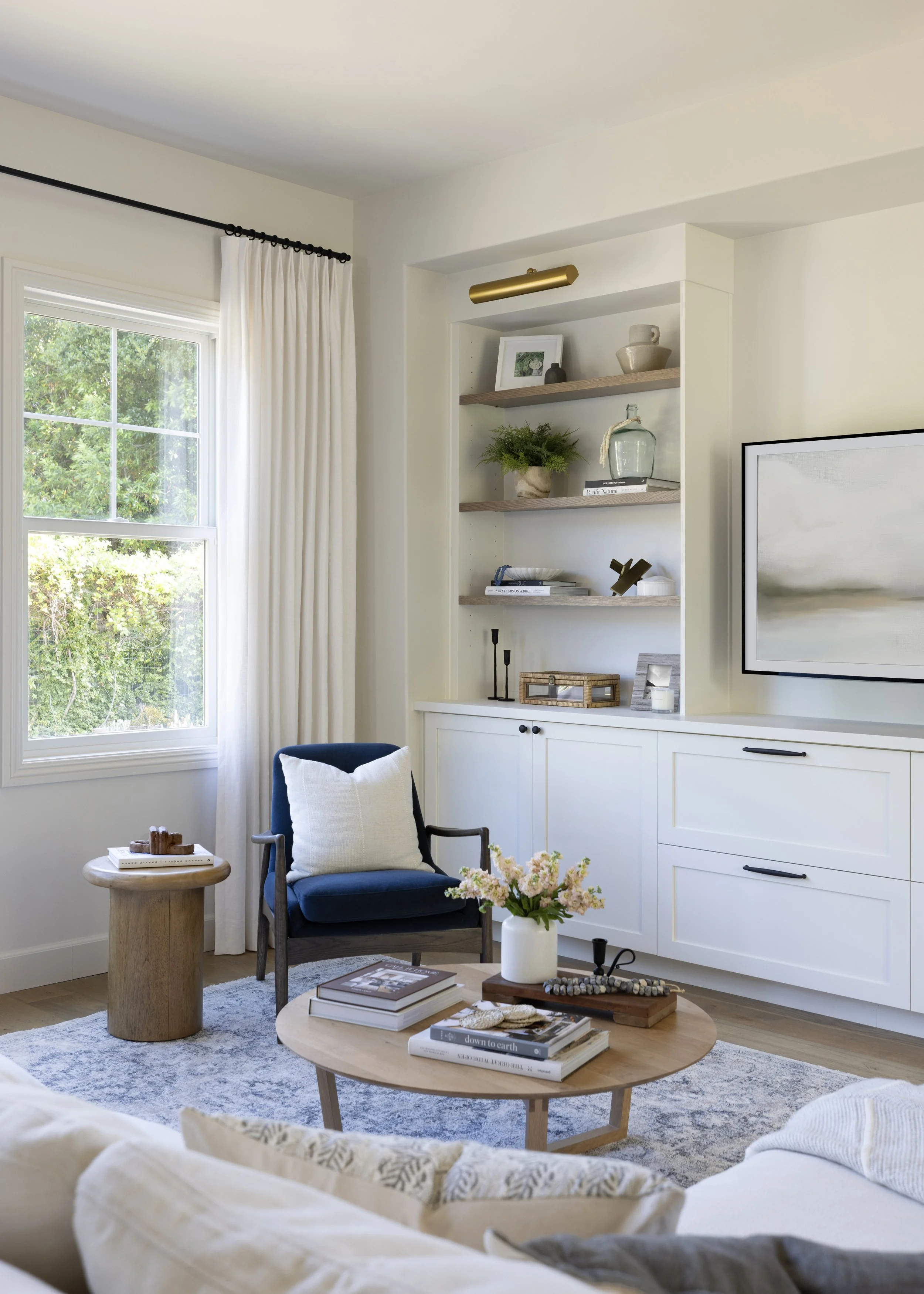 A cozy living room with a large window covered by white curtains, a blue armchair with a pillow, a small wooden side table with decorative items, a round coffee table with books and flower arrangement, a white built-in shelf with decorative objects and books, a wall-mounted television, and a soft patterned rug.
