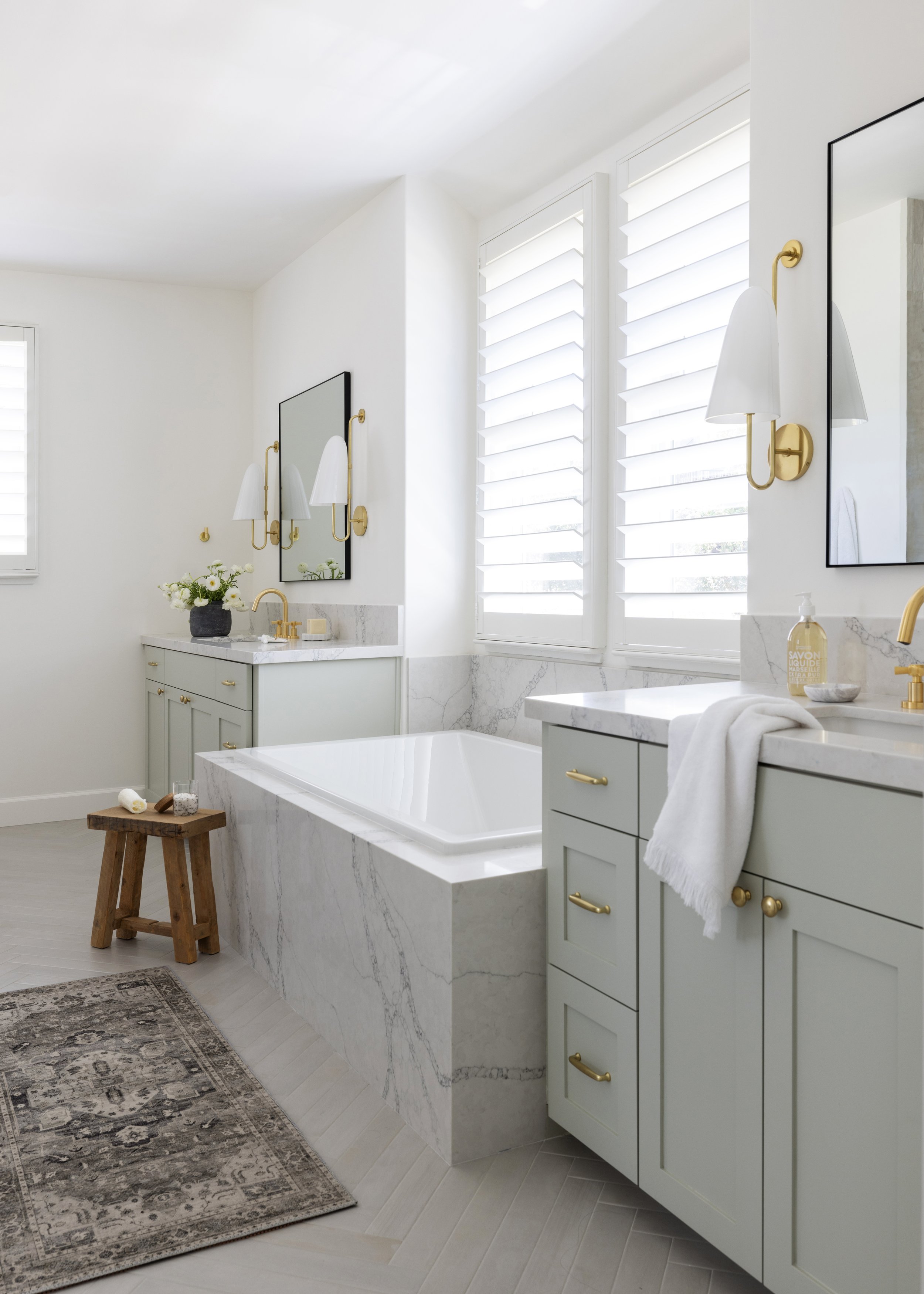 A modern, white bathroom with a white marble bathtub, a vanity with light green cabinets and gold handles, a towel hanging over the edge, gold wall sconces, a large mirror, and windows with white plantation shutters.