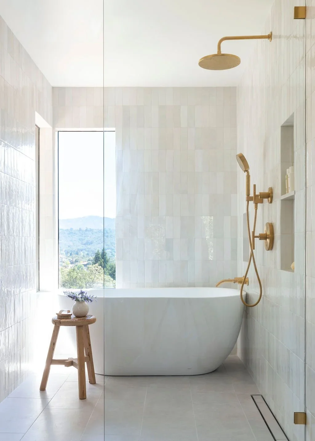 Modern bathroom with a freestanding bathtub, gold shower fixtures, a window with a view, and a small wooden stool with a vase of lavender.