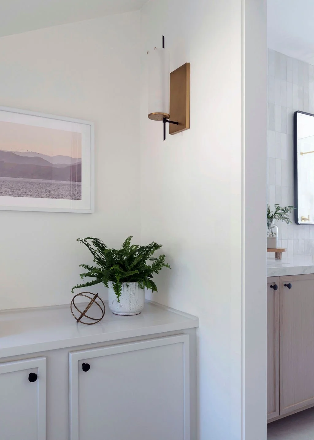 Interior view of a room with a white cabinet, a potted fern, a framed landscape photo on the wall, and a wall sconce light fixture.