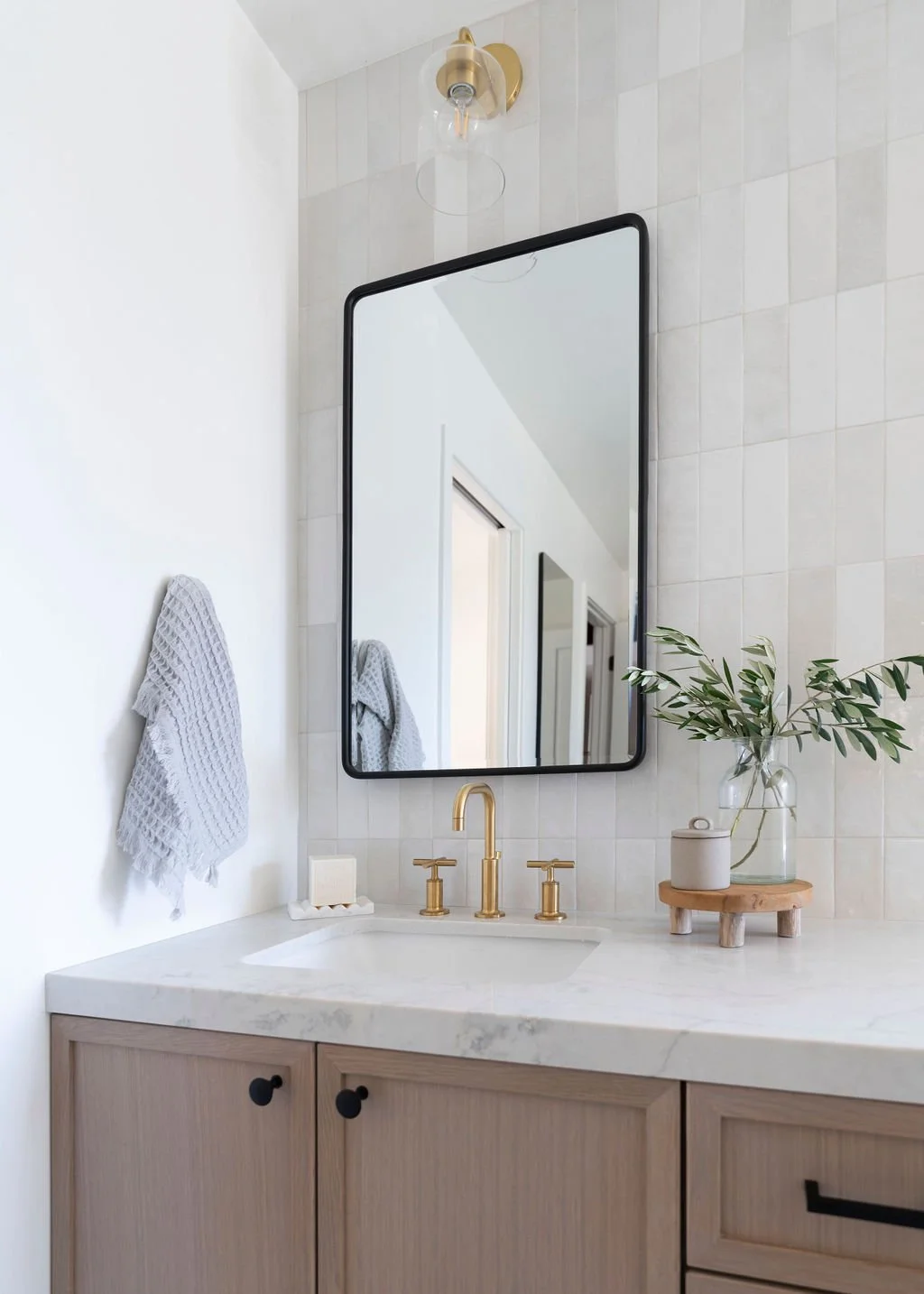A modern bathroom vanity area with a large rectangular mirror, gold faucet, light-colored cabinet, a small tray with soap, and a vase with green leafy branches on a round wooden stand. A gray textured towel hangs on the wall.
