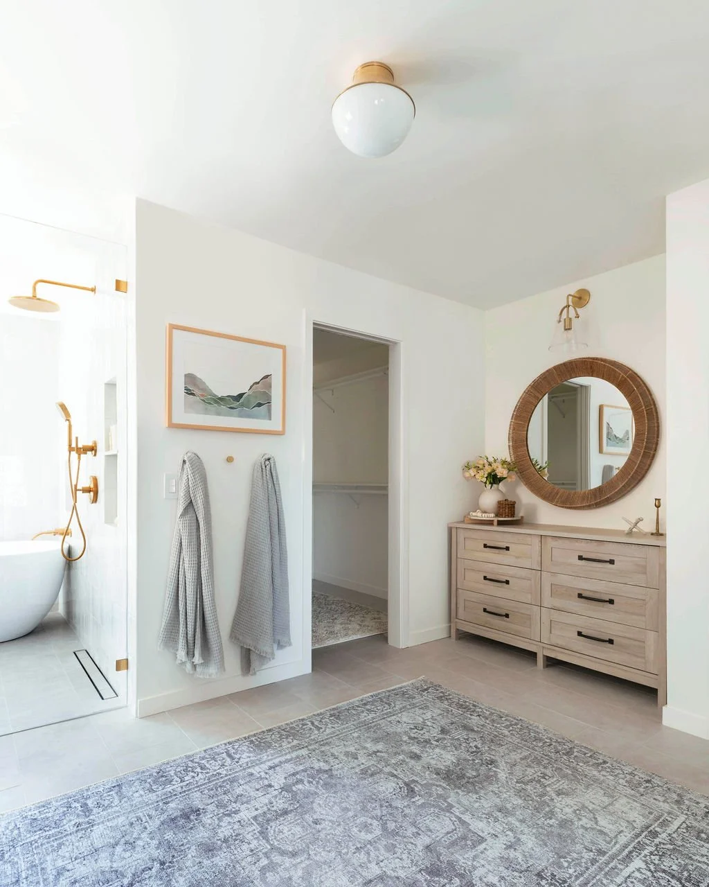 Bathroom with a wooden vanity, round mirror, floral arrangement, towels, and a walk-in shower with gold fixtures.