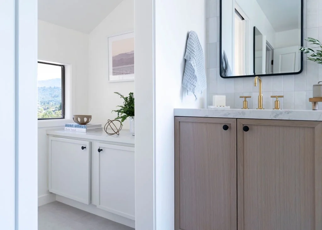Modern bathroom with a wooden vanity, gold fixtures, a large mirror, and a white towel hanging on the wall. A cabinet with decorative items and a window with a view of mountains.