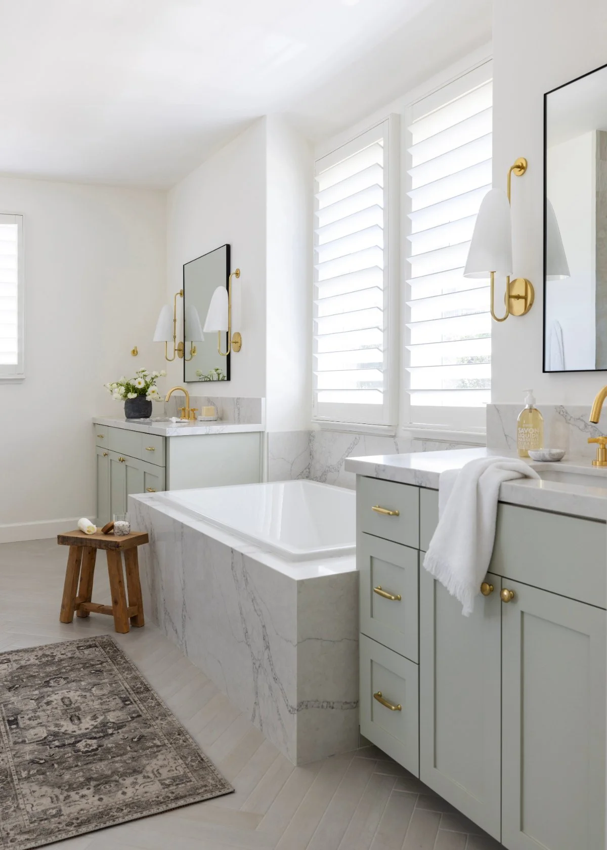 A bright, modern bathroom with white walls, large windows with shutters, a white bathtub with marble sides, light green cabinets with gold hardware, a marble countertop, a mirror, wall-mounted lamps, a small wooden stool, and decorative items like flowers and soap.