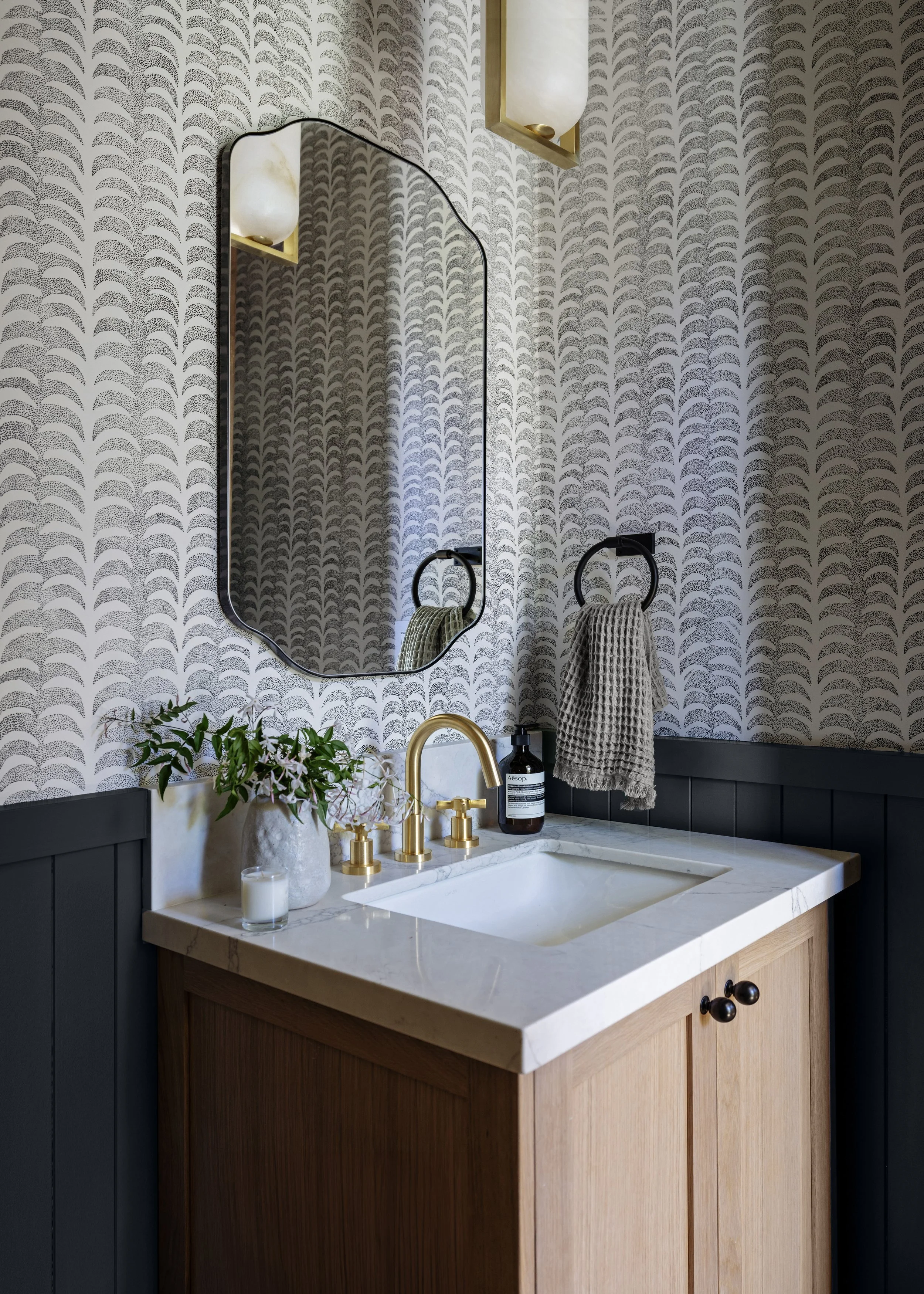 A bathroom vanity with a white marble countertop, a gold faucet, a mirror with a black frame, and wall-mounted light fixtures. The wall has patterned wallpaper, and there are decorative items including a vase with greenery, a candle, and a bottle of 