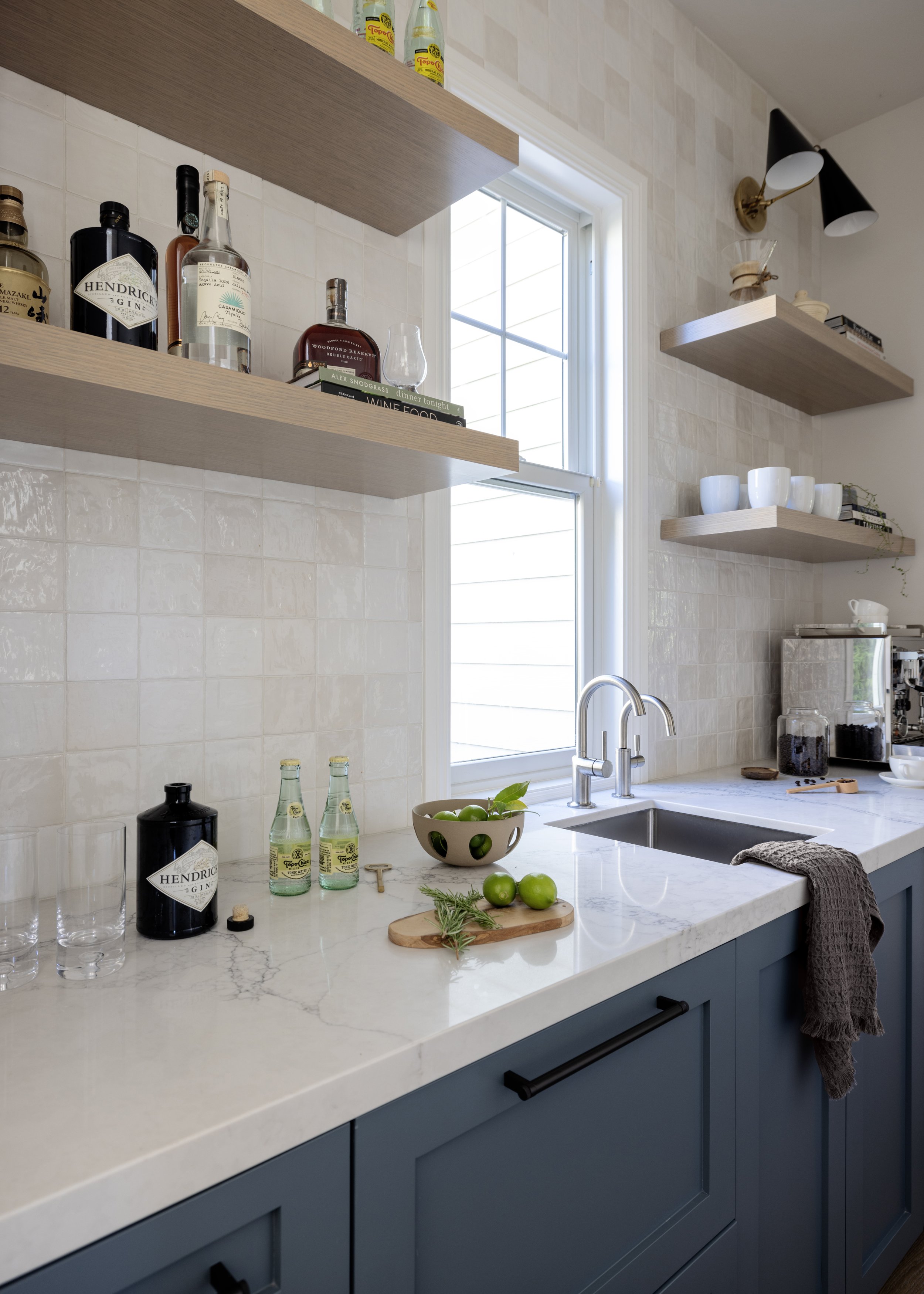 A modern kitchen with blue cabinets, a white marble countertop, and open wooden shelves. Items on the shelves include bottles, cups, and books. There is a window above the sink with a view outside. On the countertop, there are green apples, a bowl, h