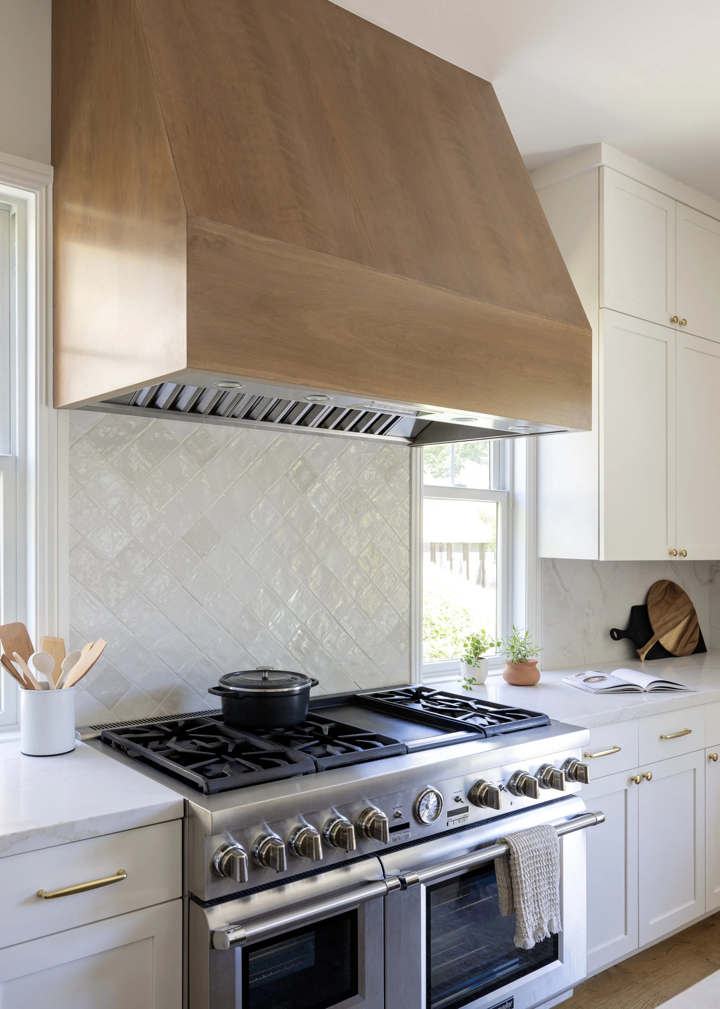 A modern kitchen with a white countertop, white cabinets with gold handles, a stainless steel stove and oven, a wooden range hood, a window with plants on the sill, and kitchen utensils in a white holder.