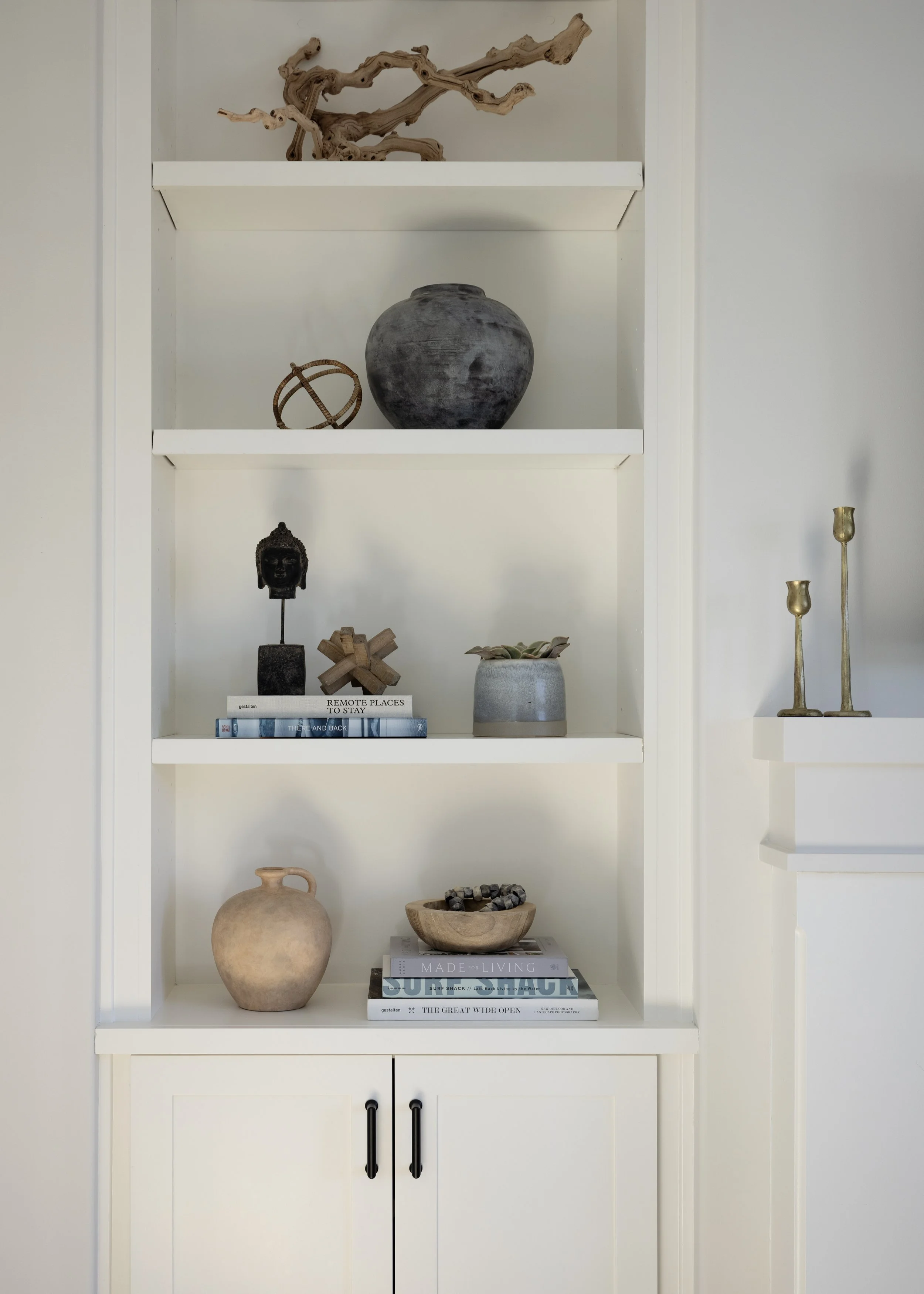 A white bookshelf with three shelves holding decorative items, including a driftwood sculpture, a gray ceramic vase, a small sphere sculpture, a black Buddha head, a wooden cross, plants, and books.