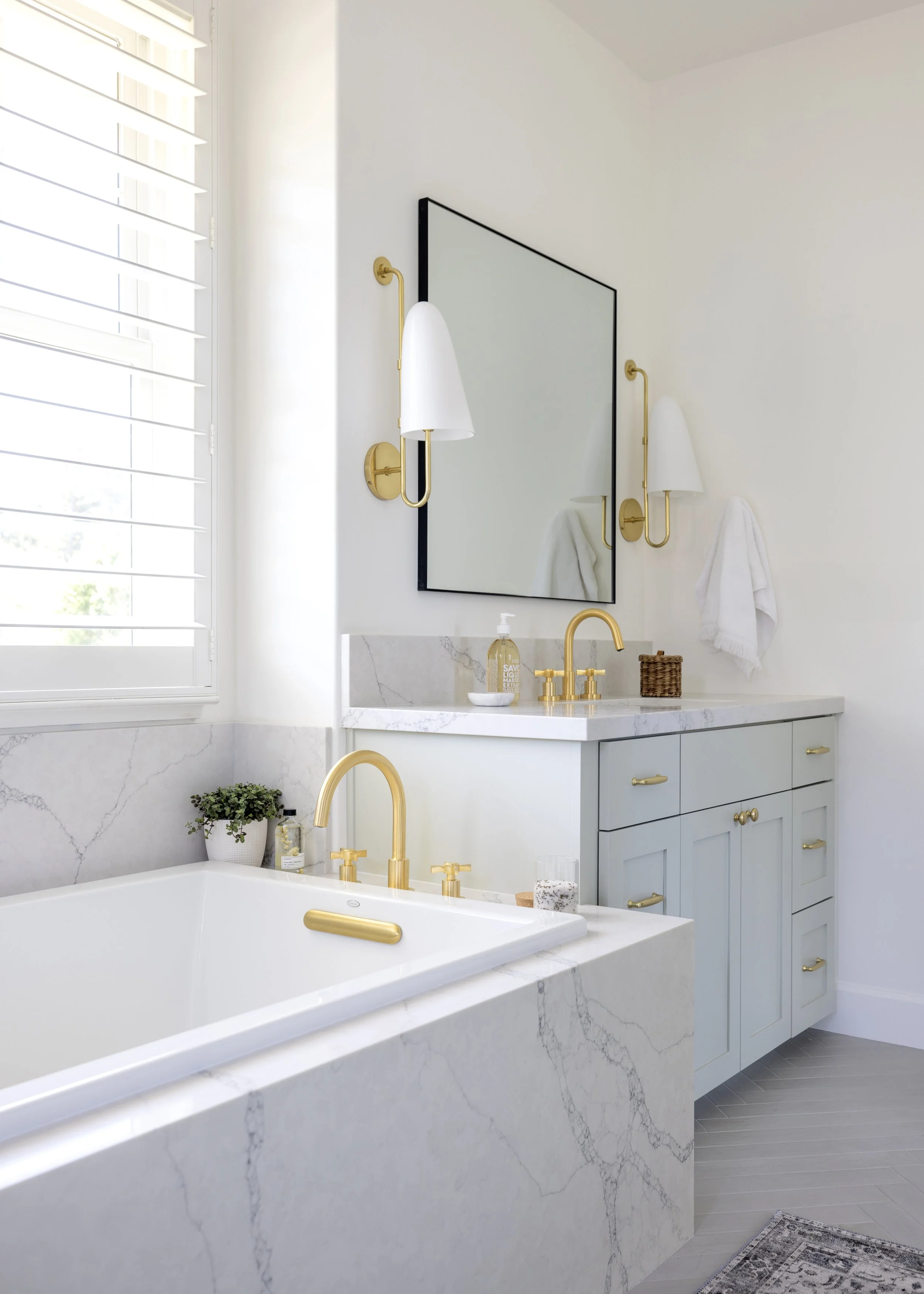 A modern bathroom with white marble surfaces, a large mirror, gold fixtures, a white bathtub, a potted plant, and white walls with a window with shutters.