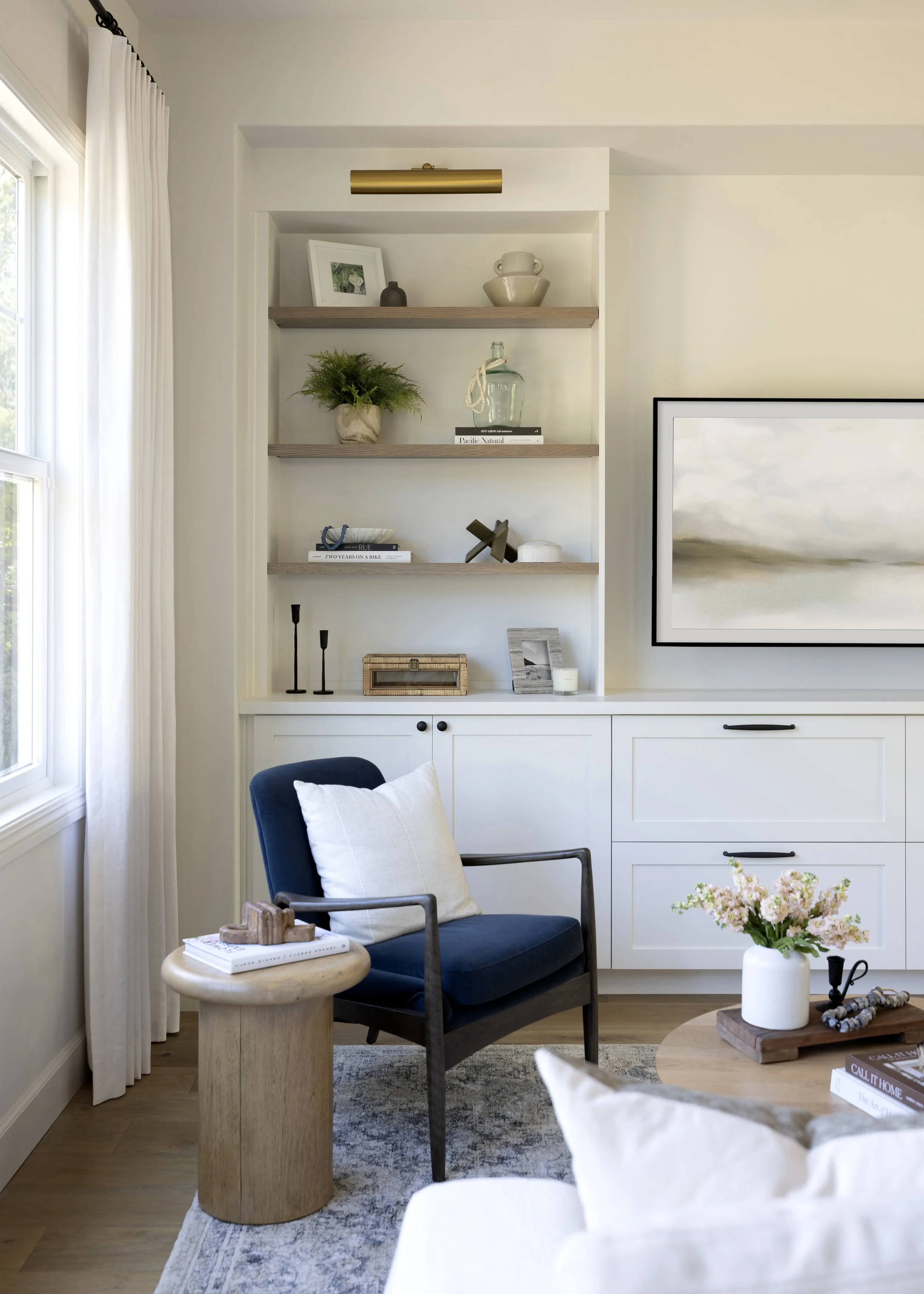 A cozy living room corner with a navy blue armchair, beige cushion, wooden side table with books and decorative items, a white built-in cabinet with open shelving decorated with plants, vases, and framed pictures, a large framed landscape photograph 