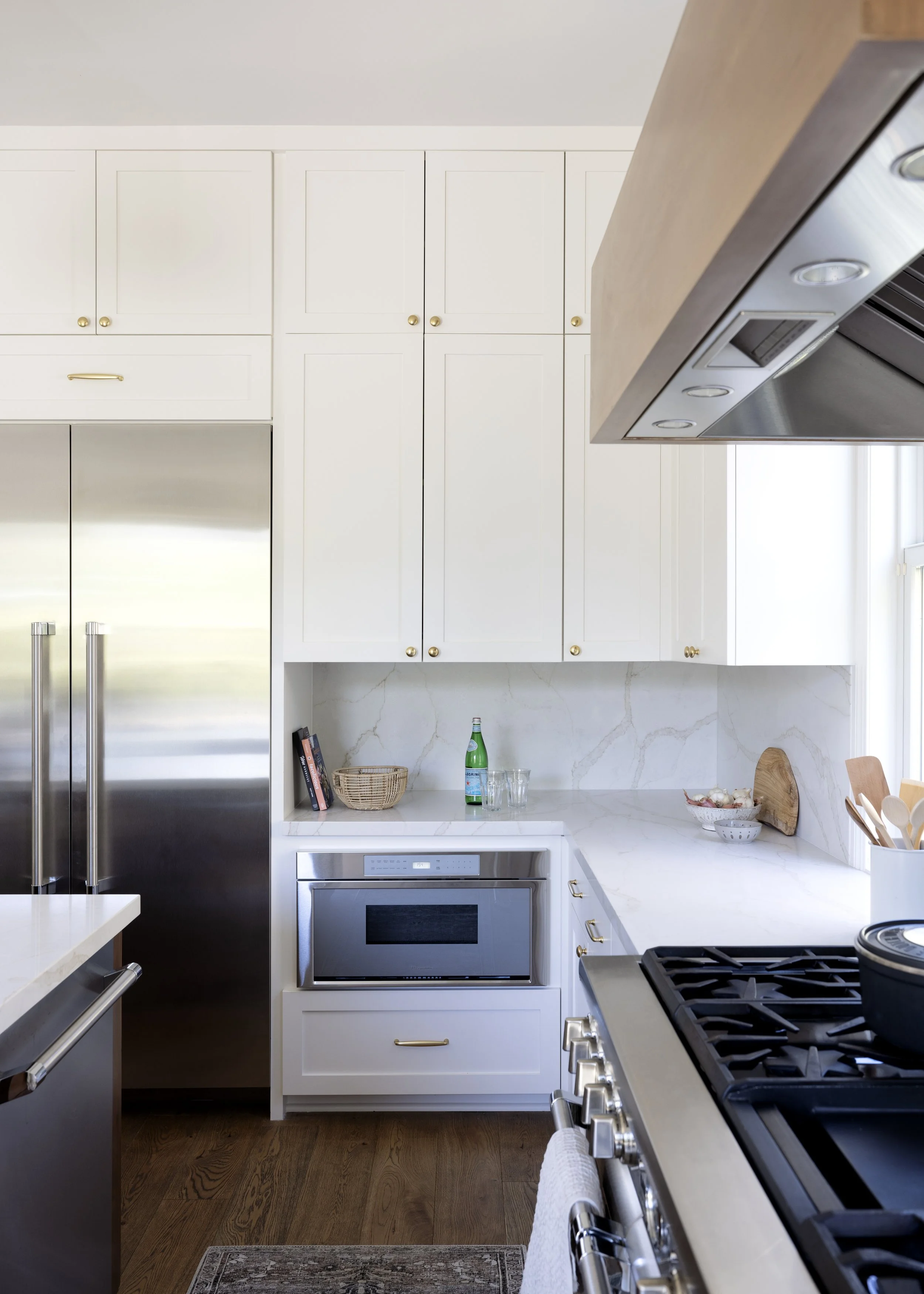 Modern kitchen with white cabinetry, stainless steel refrigerator, microwave oven, gas stove, wood flooring, and a white marble countertop with some kitchen items and a small patterned rug.