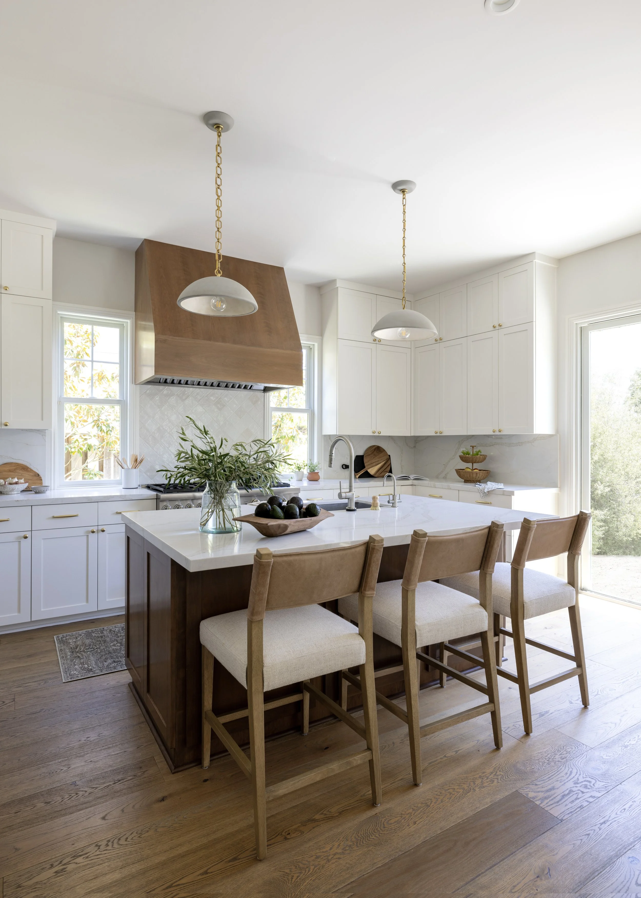 A modern kitchen with a white marble island, four beige chairs, white cabinets, a wooden ceiling hood over the stove, and large windows bringing in natural light.