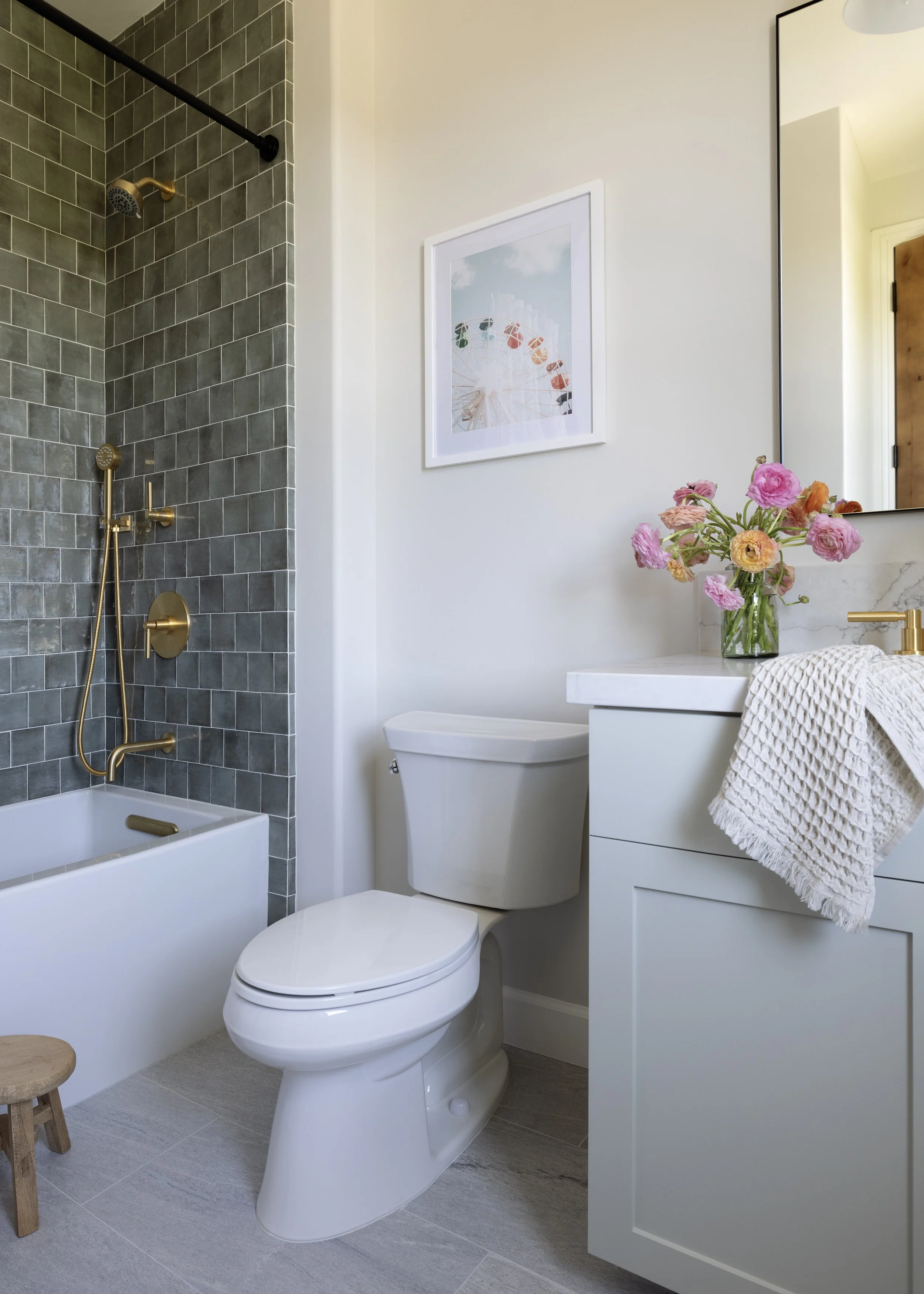 A modern bathroom with a white toilet, gray tile shower with brass fixtures, a framed picture of a Ferris wheel, a white vanity with a gold faucet, a mirror, a vase of pink and orange flowers, and a beige towel.