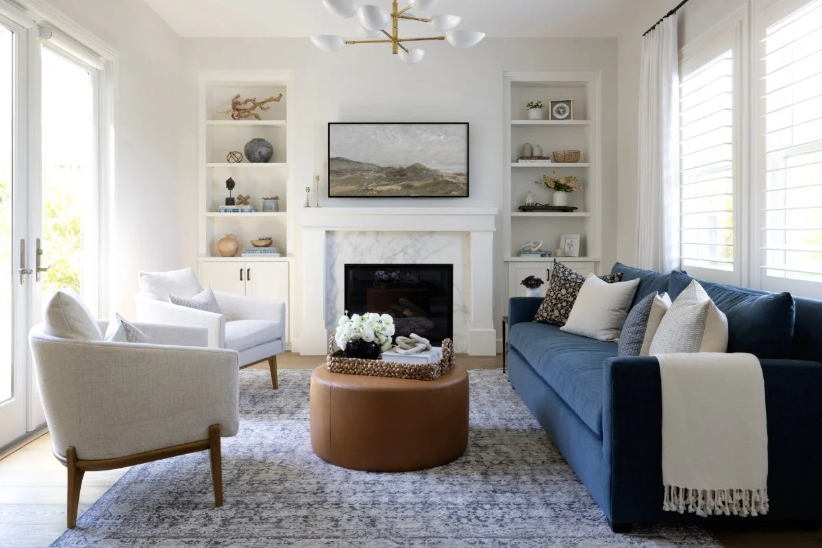Living room with white walls, a fireplace, a blue sofa, white armchairs, and built-in shelves filled with decorative items. Large windows with white blinds provide natural light.