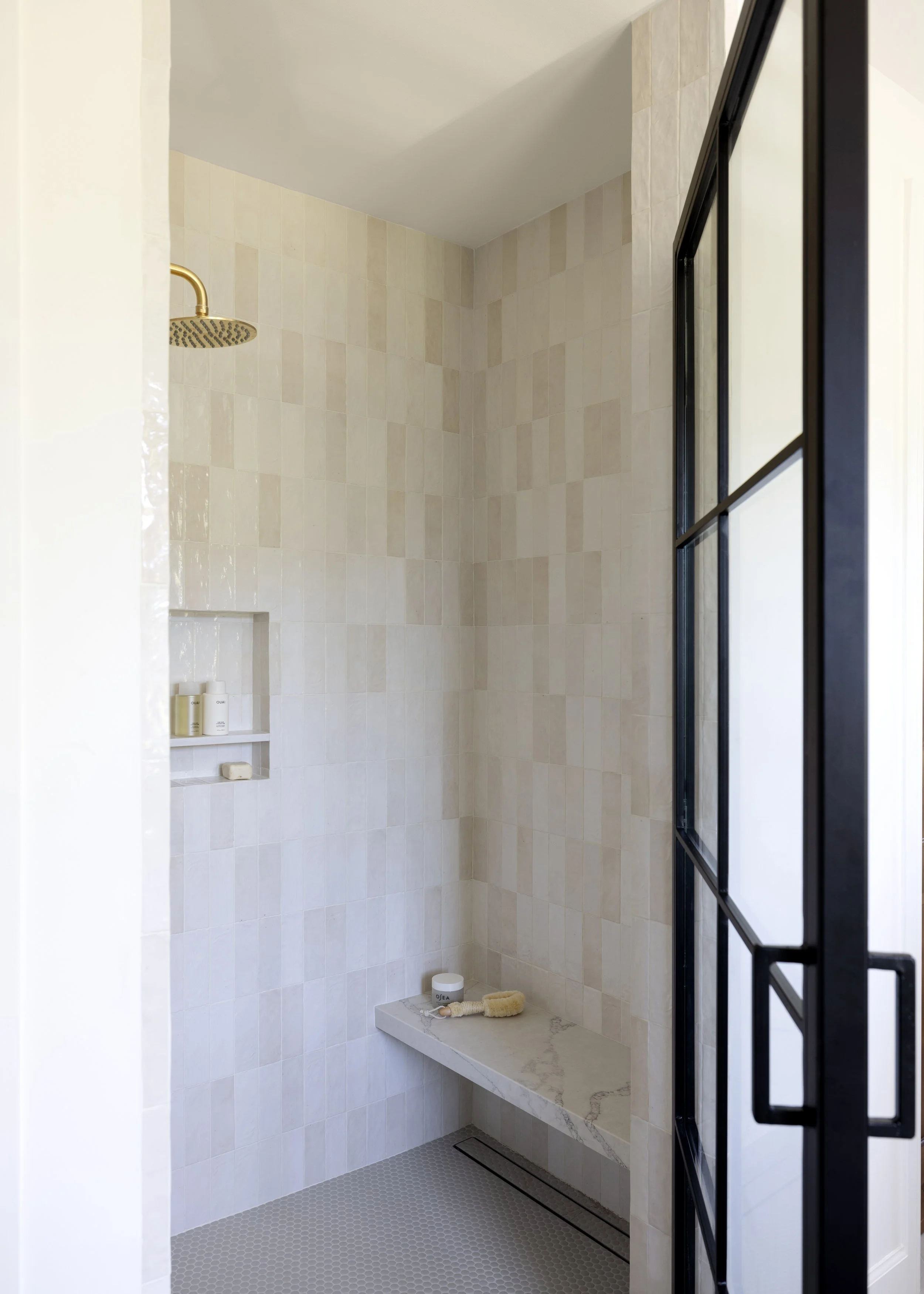 Modern shower with beige tile walls, a rainfall showerhead, a marble bench with bath products, and a black-framed glass door.