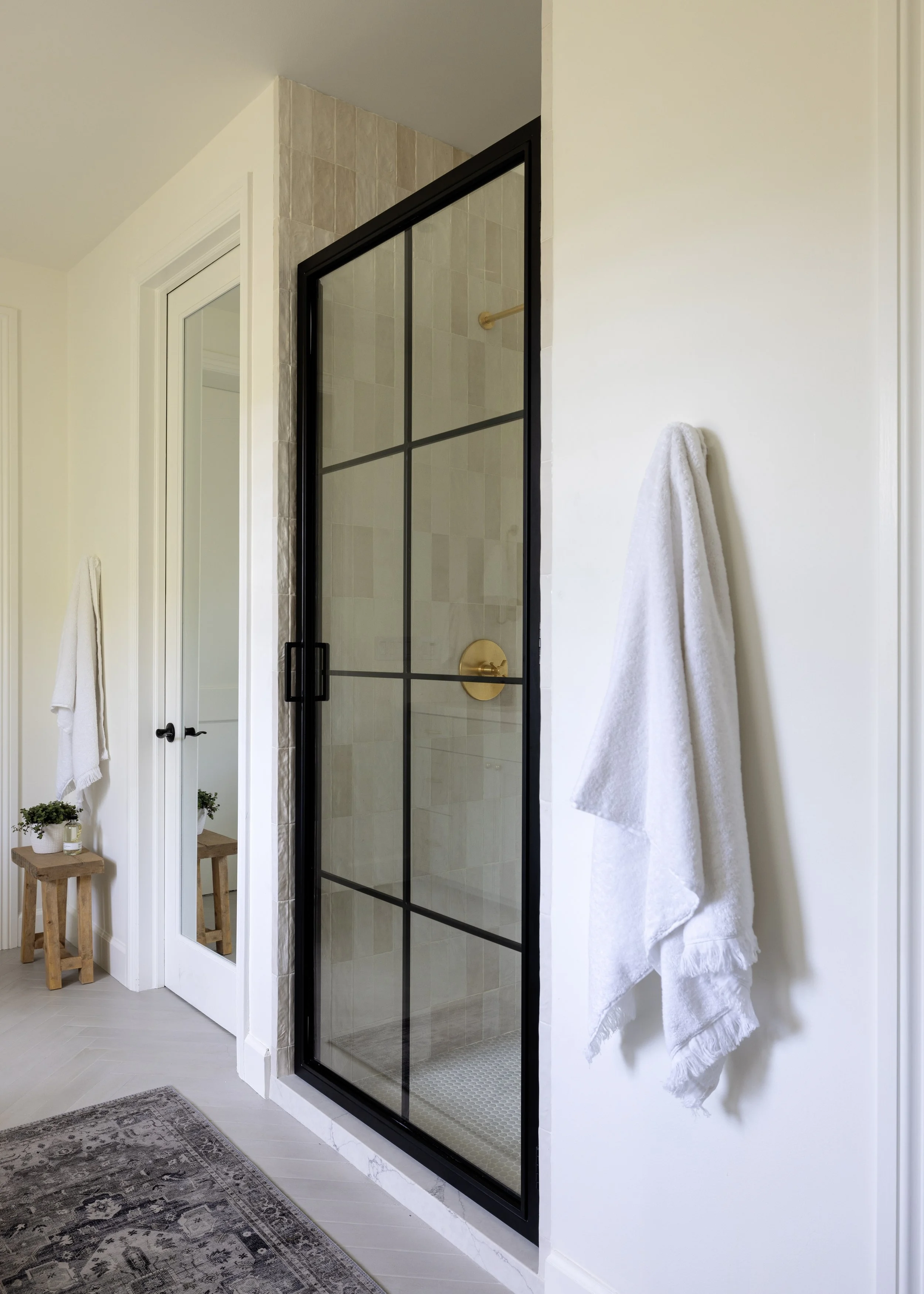 A bathroom with a glass shower enclosure with black framing, a gold shower handle, and beige tiled shower walls. A white towel hangs on a hook on the wall, and there is a wooden stool with small plants on top and a white towel underneath. Part of a m