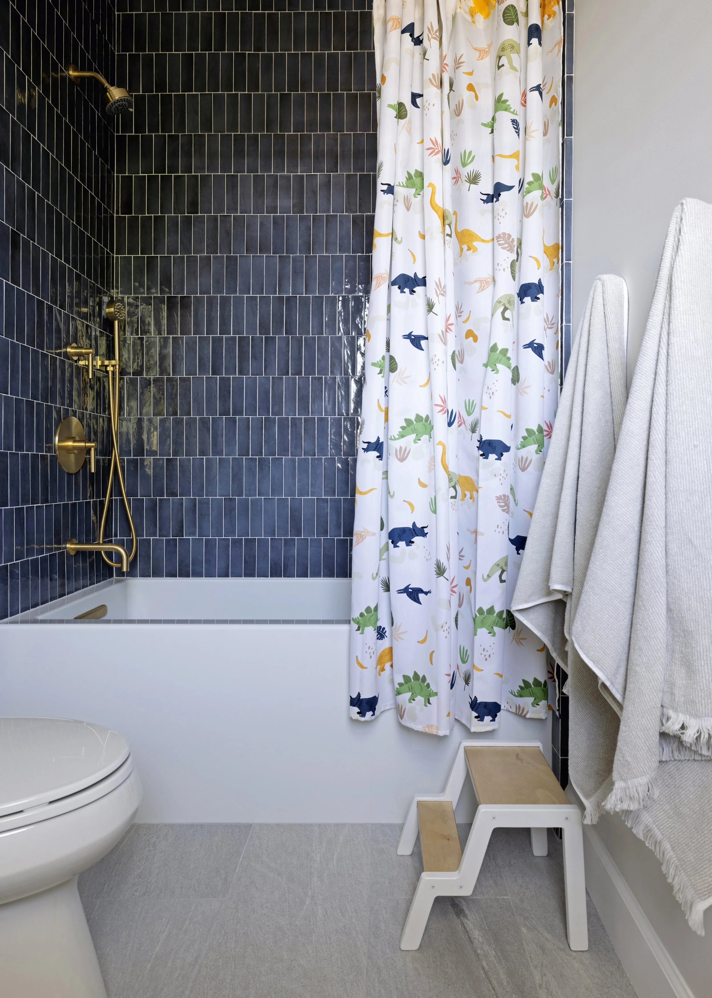 Modern bathroom with black tiled shower area, yellow-golden fixtures, and a colorful dinosaur print shower curtain. White towels hang on a rack, with a small wooden step stool and a white step stool in the corner. The bathroom has light gray flooring