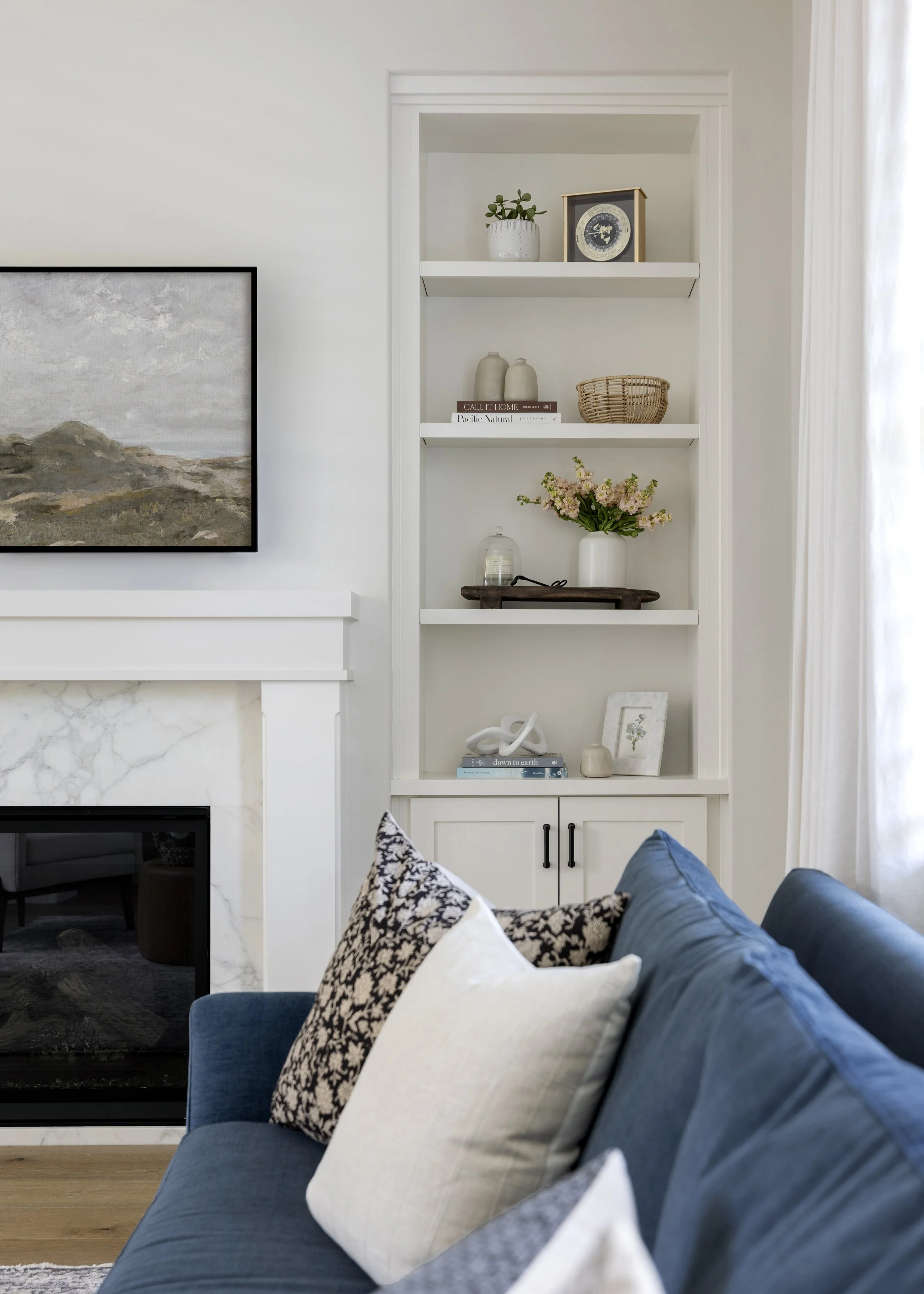 Living room with a blue sofa and pillows, a white marble fireplace, and a built-in white shelf with decorative items including plants, vases, books, and framed art, with a large window with curtains on the right side.