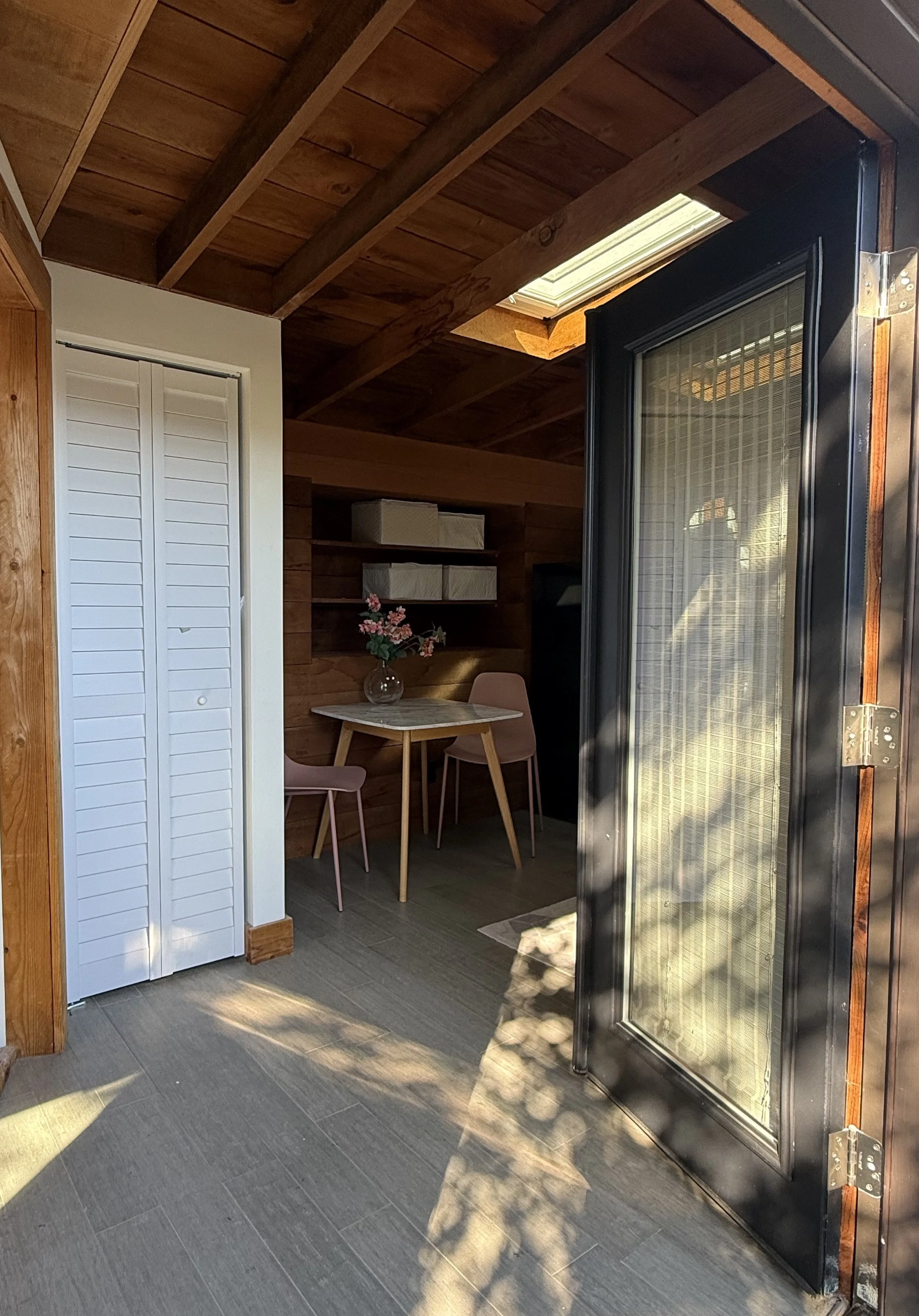 A small indoor patio or balcony area with a table, two pink chairs, shelving with boxes, a vase with pink flowers, a sliding glass door, and a wooden ceiling with a skylight.