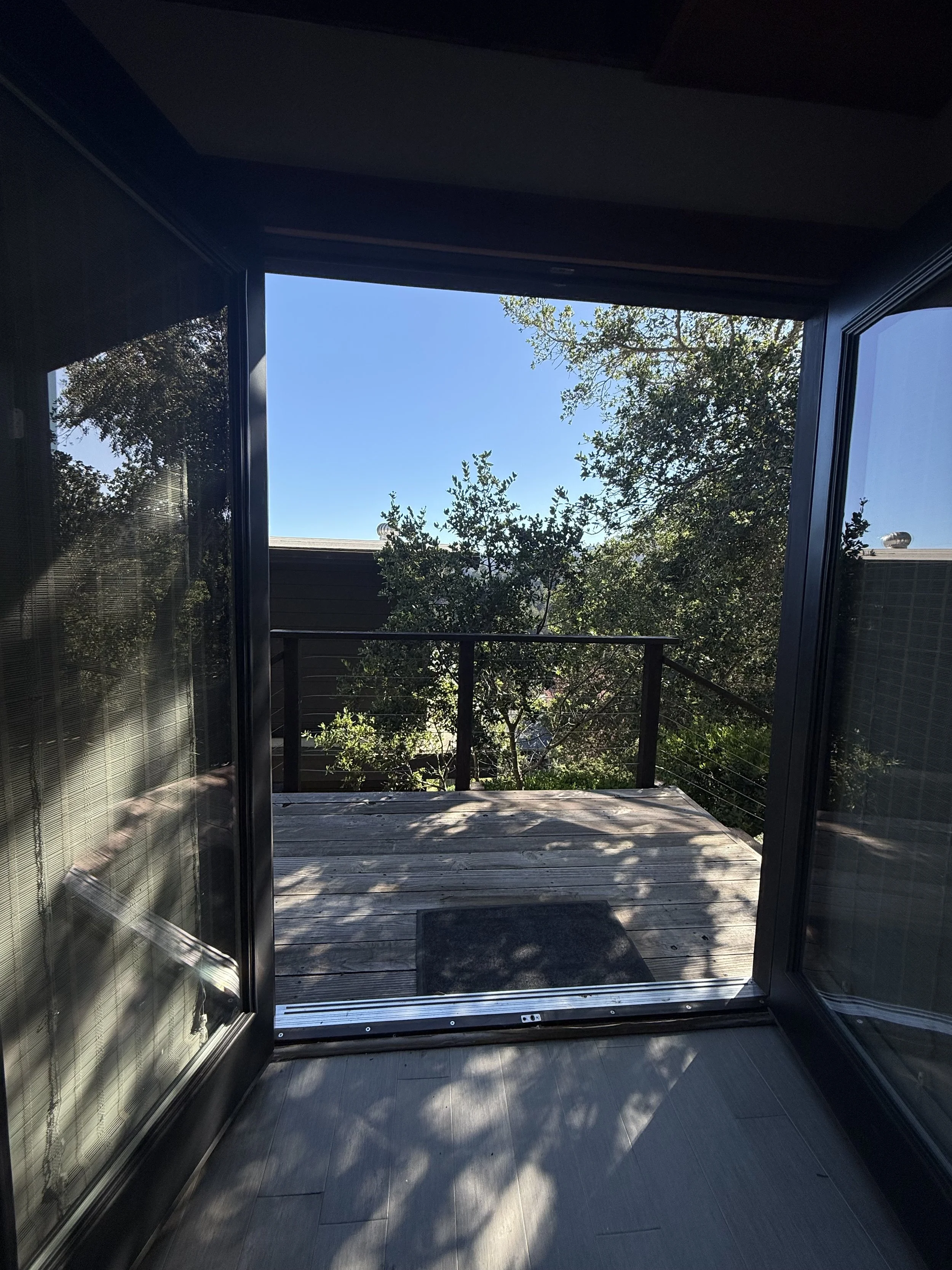 Open glass door leading to a small wooden deck with a black metal railing, overlooking trees and a clear blue sky.