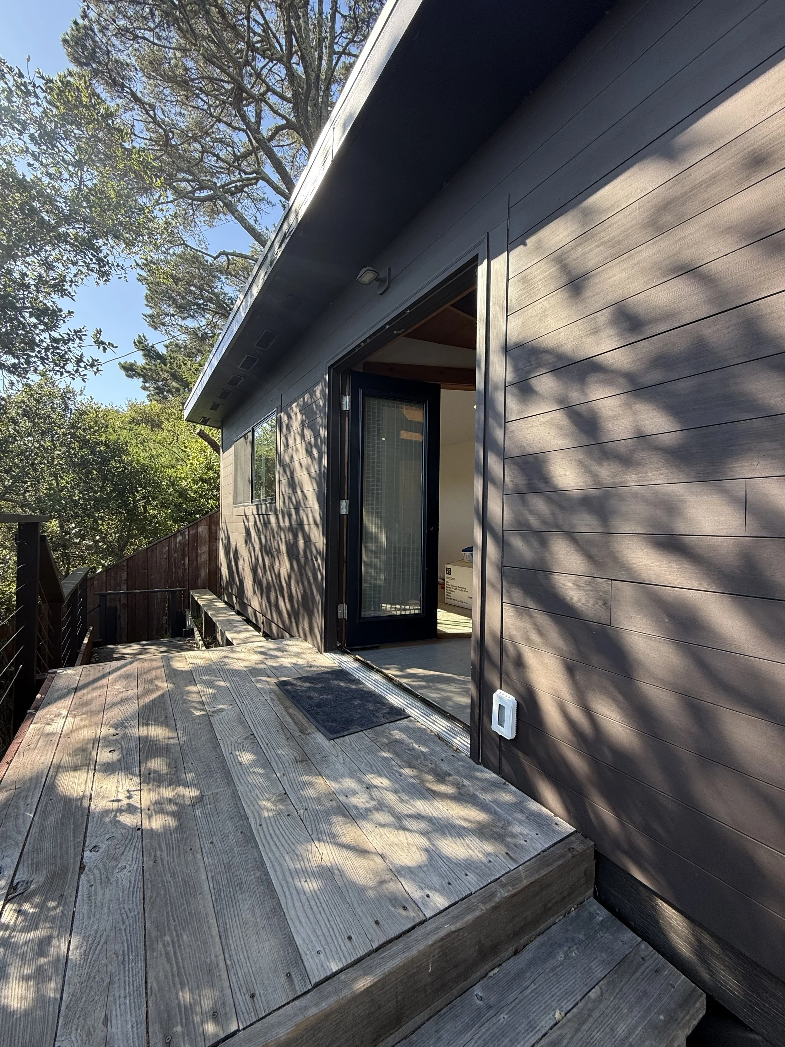 Backyard patio with wooden deck, sliding glass door, shadow of tree branches on the wall, trees and blue sky in the background.