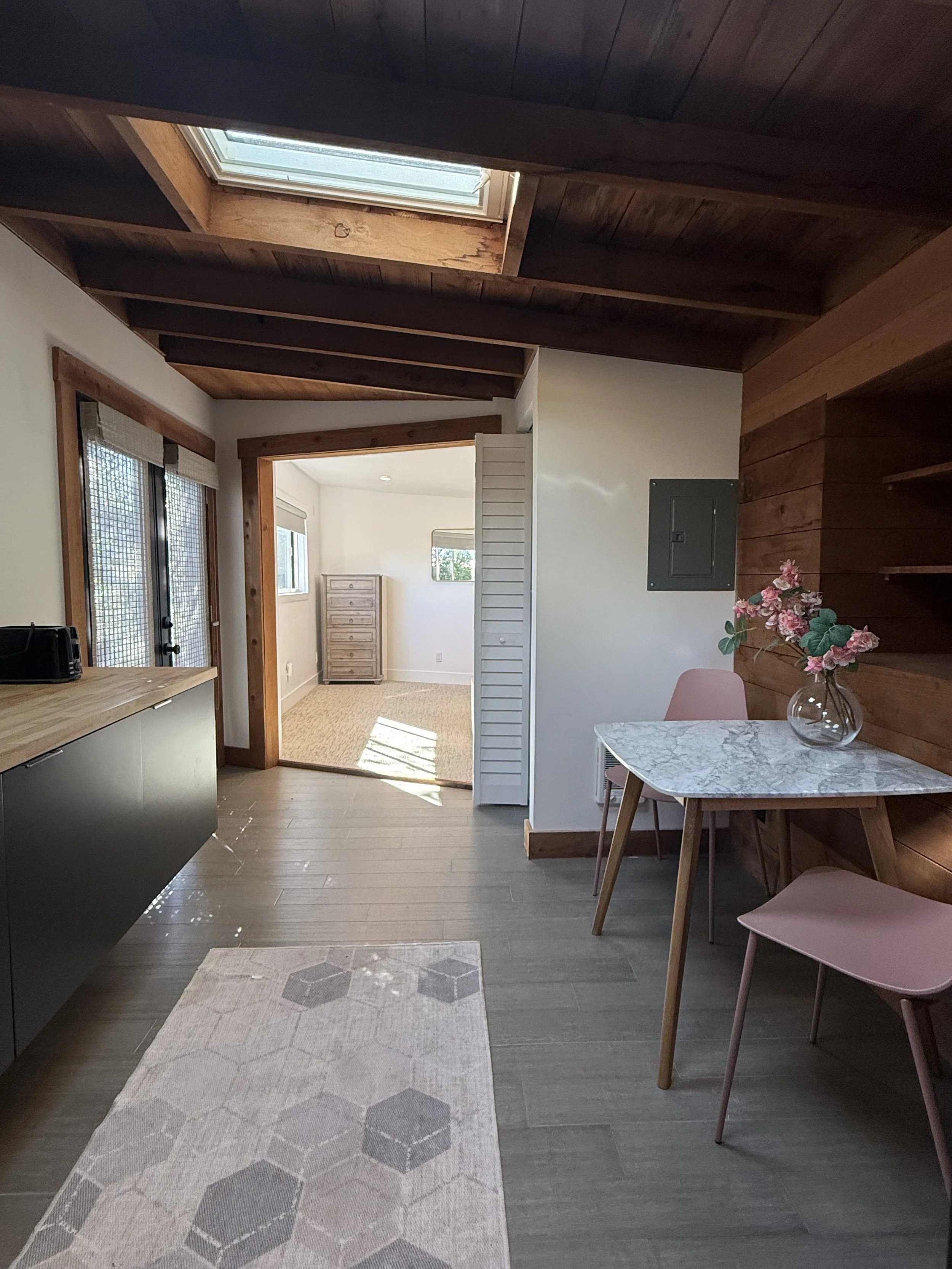 Interior of a room with wood-paneled ceiling, a skylight, a gray and white hexagon-patterned rug, a marble-top table with pink chairs, and a flower vase with pink flowers.