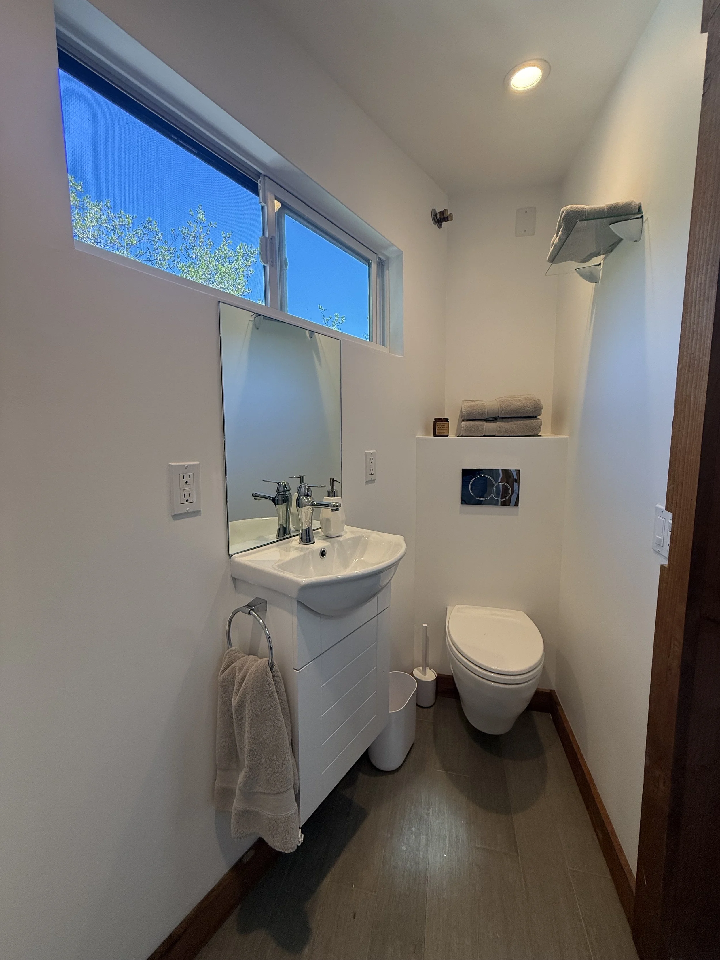 A small bathroom with a white sink, a mirror, a toilet, a window with blue sky outside, towels on a shelf, and a bottle of liquid soap on the sink.