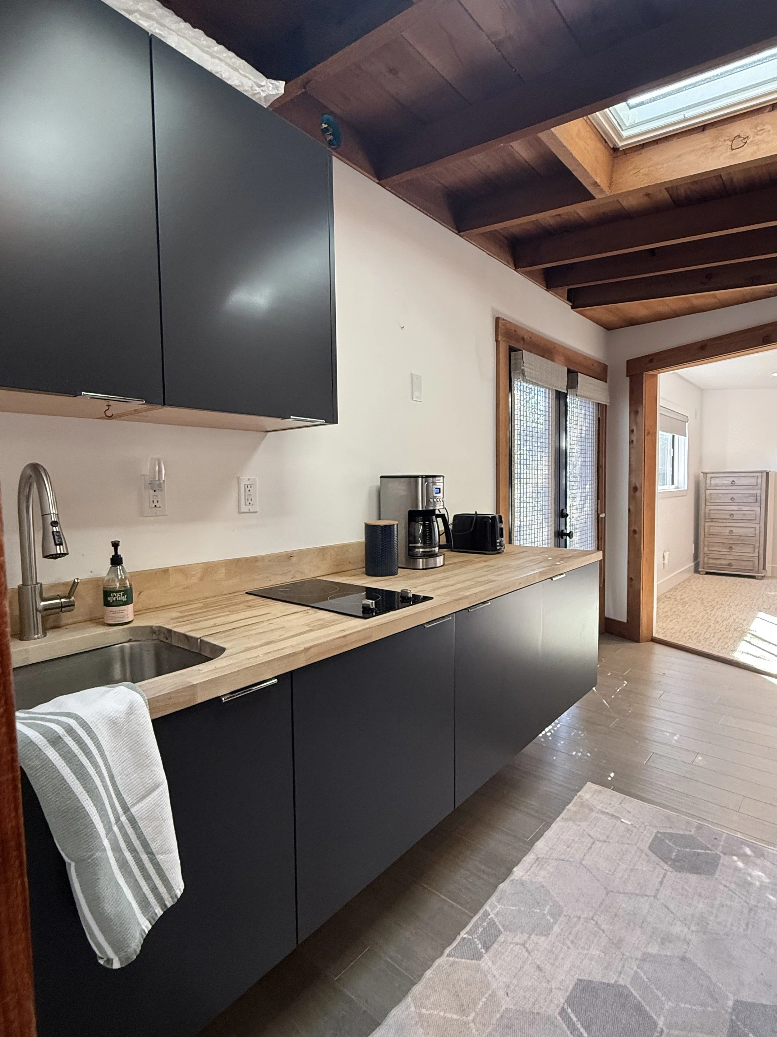 Modern kitchen with black cabinets, wooden countertop, stainless steel sink, coffee maker, and toaster. There are two windows with wooden frames and a skylight in the ceiling, with a beige rug on the wooden floor.