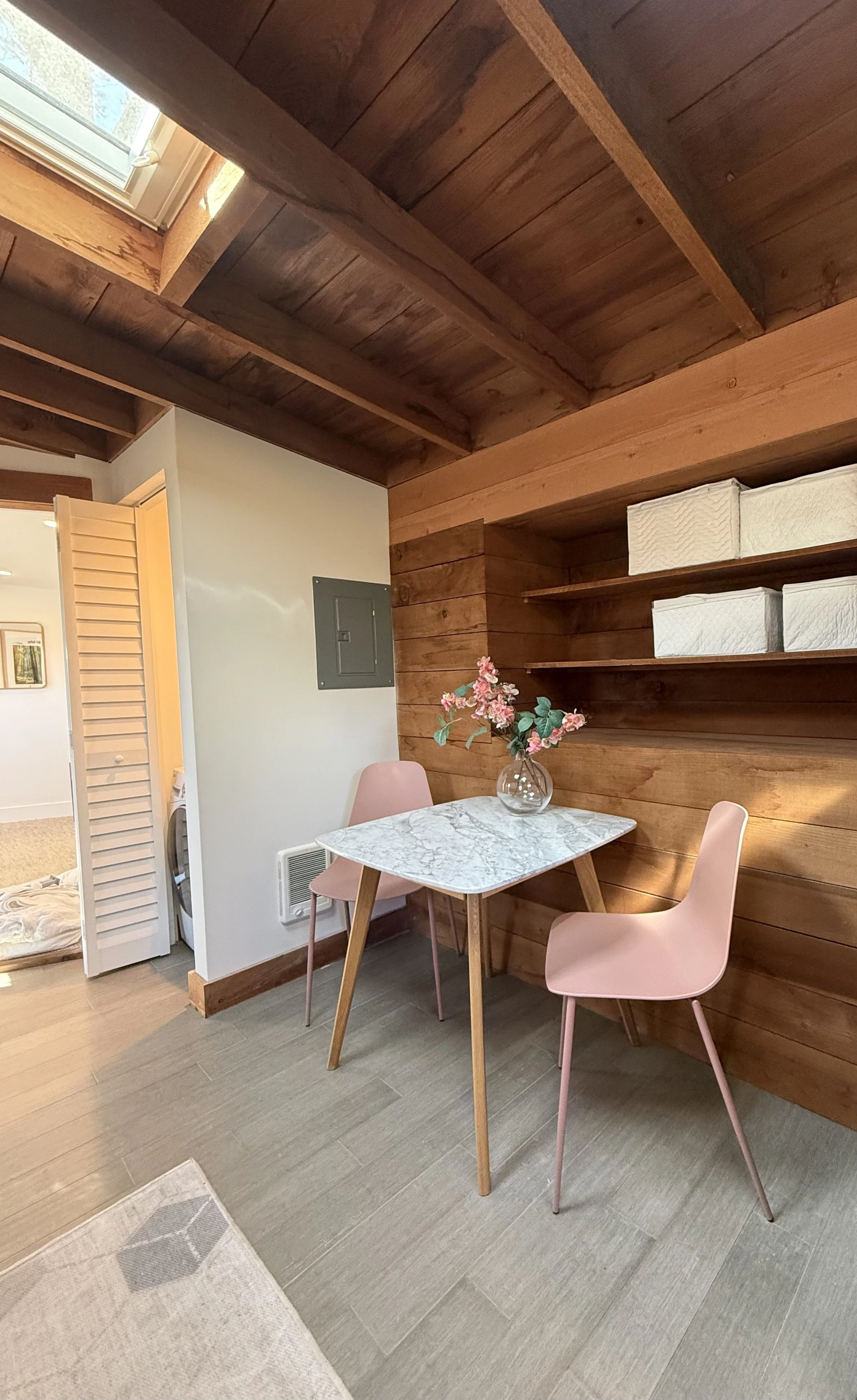 A cozy dining area with a small marble top table and two pink chairs. A vase with pink flowers is on the table, with a wooden paneled wall behind. Shelves with white boxes are mounted on the wall. There is a window in the ceiling allowing natural lig