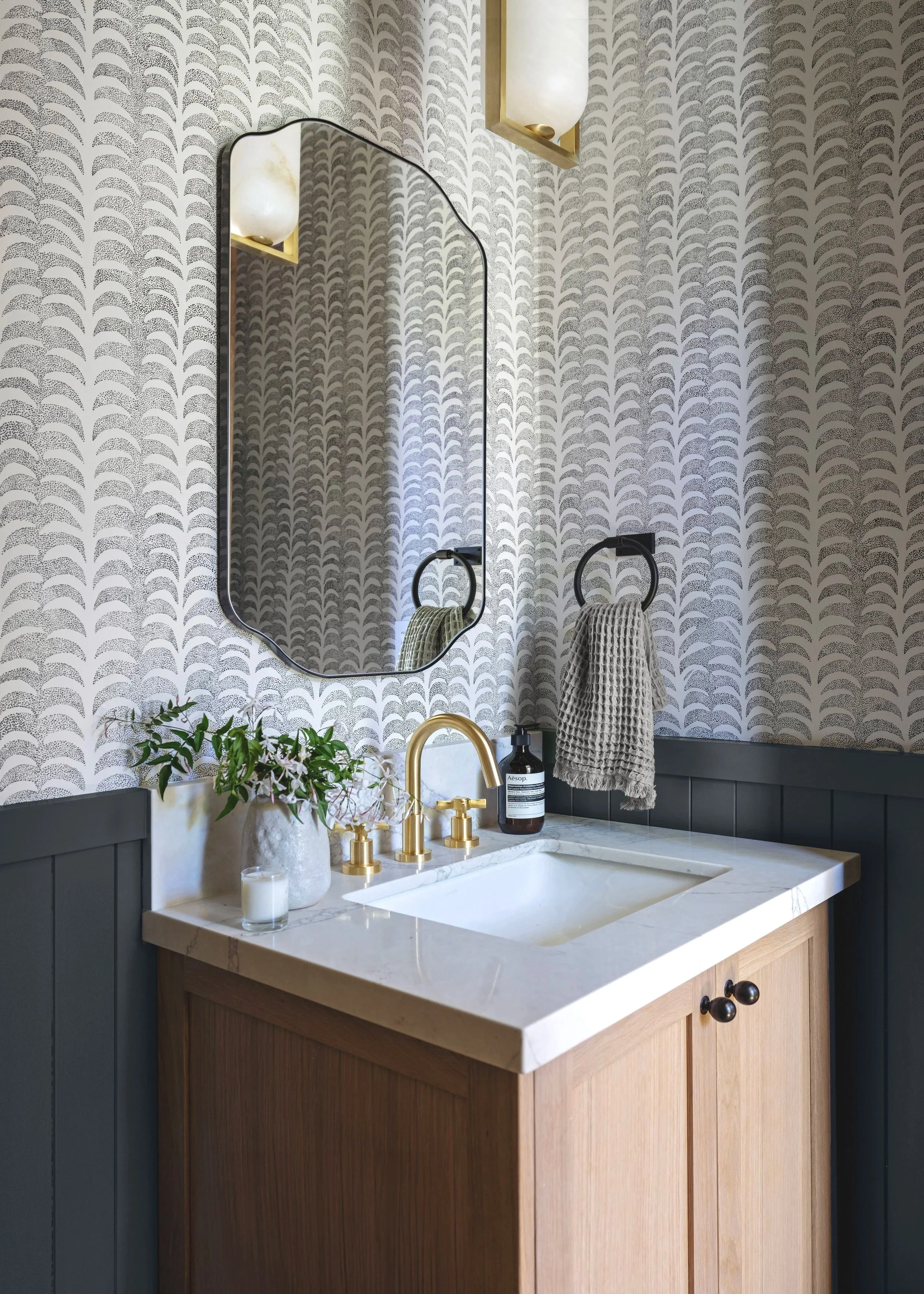 Bathroom vanity with a white marble top, gold faucet, and black cabinetry. A round mirror with a black frame hangs above the sink. On the left side of the vanity, there is a white vase with green plants and white flowers. On the right side, there is a black soap dispenser and a brown bottle. A beige towel with a textured pattern hangs on a black ring. The wall behind has gray and white patterned wallpaper and two wall-mounted light fixtures with white glass shades.