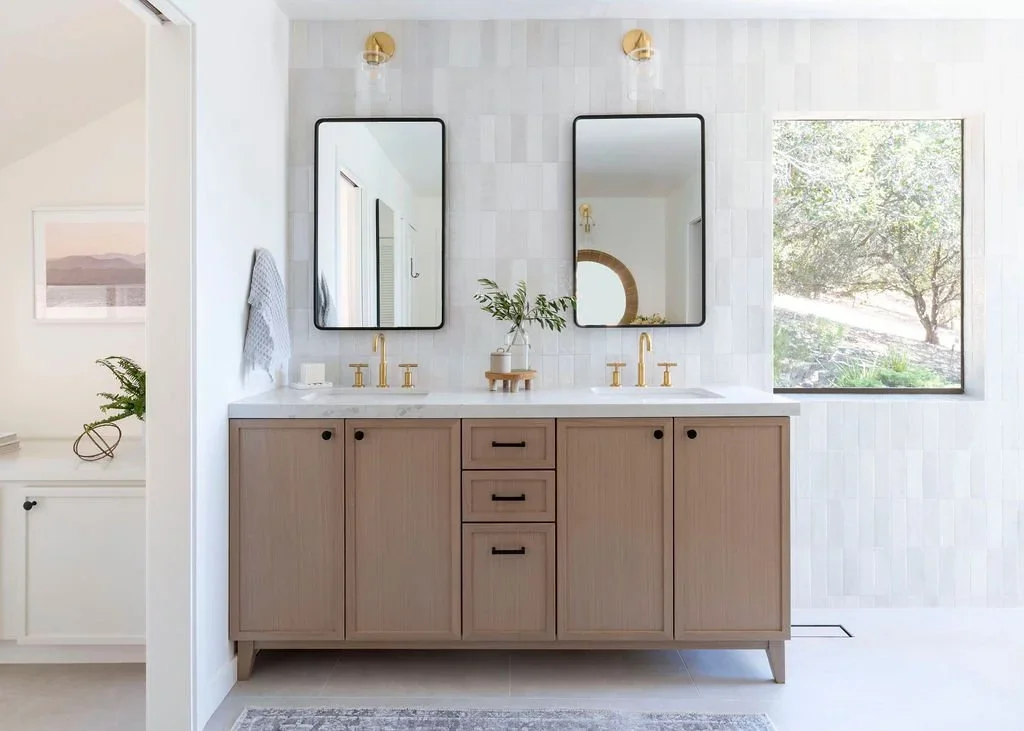 Modern bathroom with dual sinks, light wood vanity, two rectangular wall mirrors, gold fixtures, a large window showing green trees, and minimal decor.