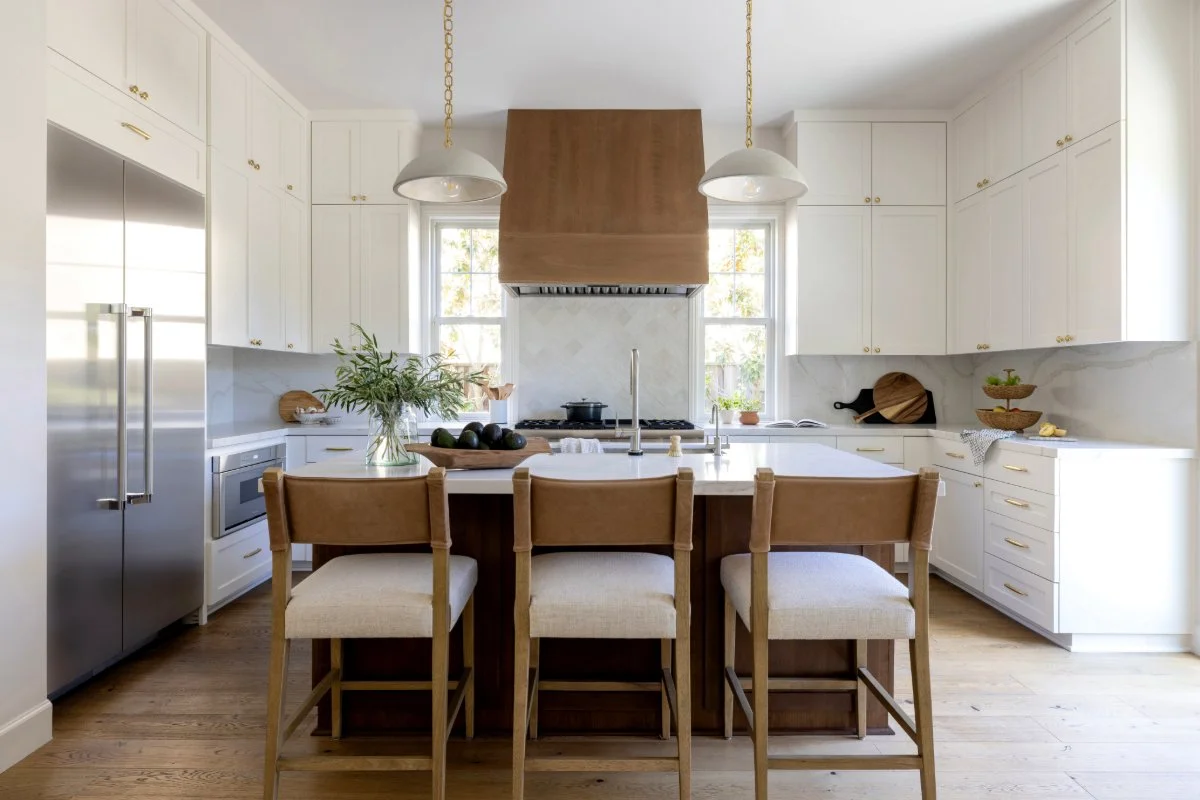 Modern kitchen with white cabinets, a central island, stainless steel refrigerator, and white marble backsplash, featuring a window behind the stove, pendant lights above the island, and wooden accents.