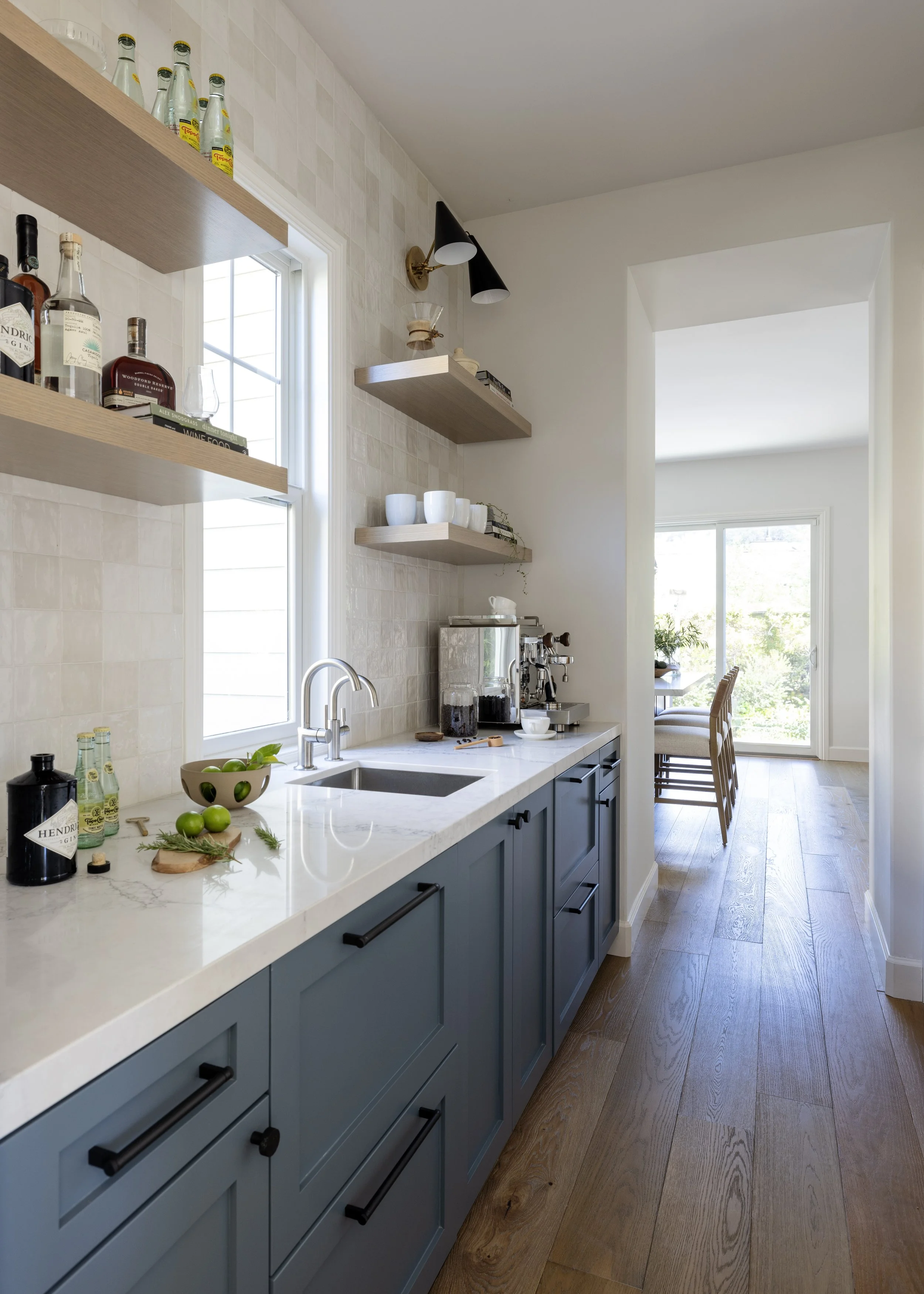 Modern kitchen with white countertop, blue cabinets, open wooden shelves, and a window. Items include bottles, cups, a bowl of green limes, and kitchen appliances, with sunlight streaming in.