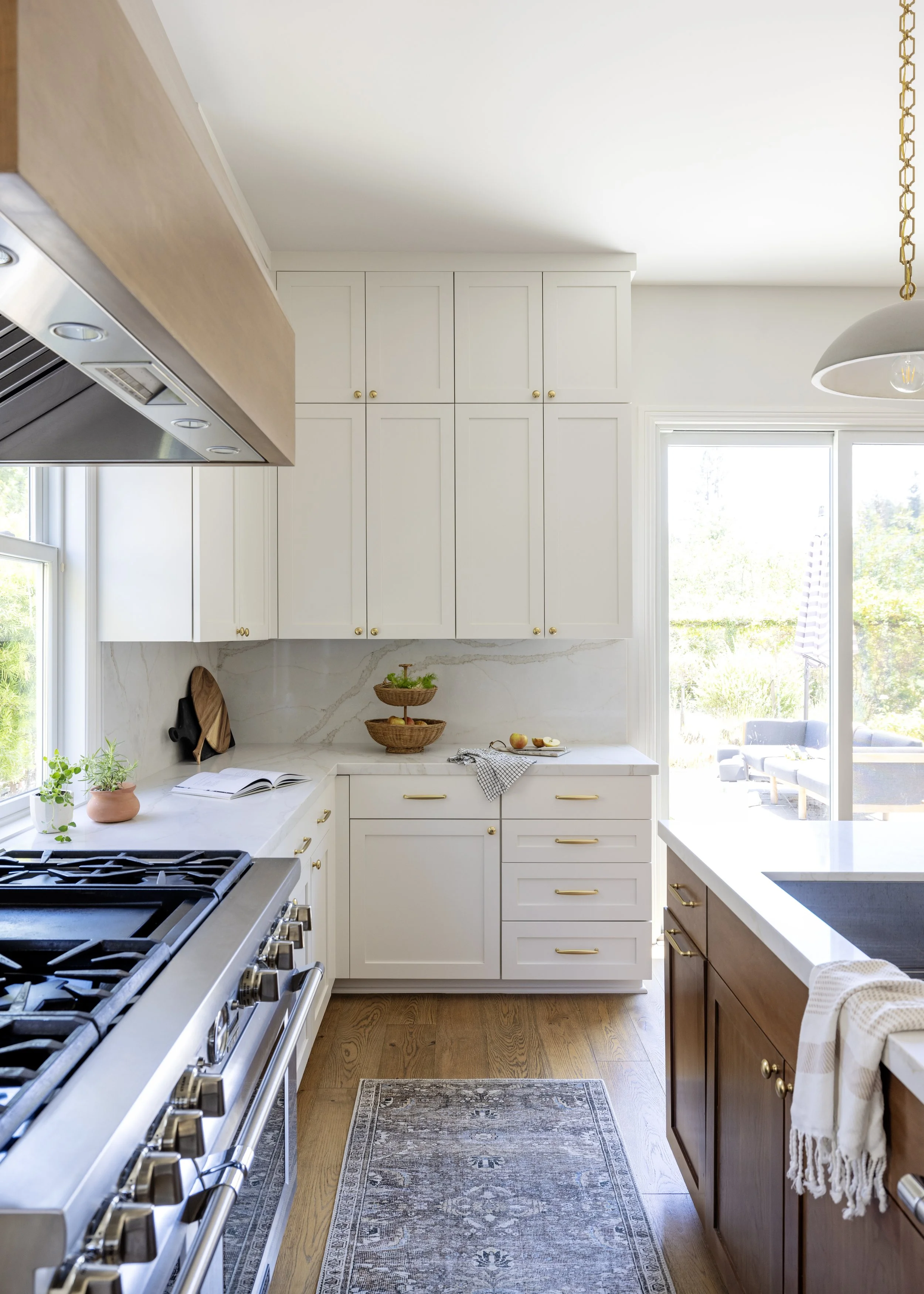 A bright kitchen with white cabinets, a gas stove, a gray oven, and a large window with a view of greenery outside. There is a kitchen island with a sink, a patterned area rug, and decorative baskets with fruit on the counter.
