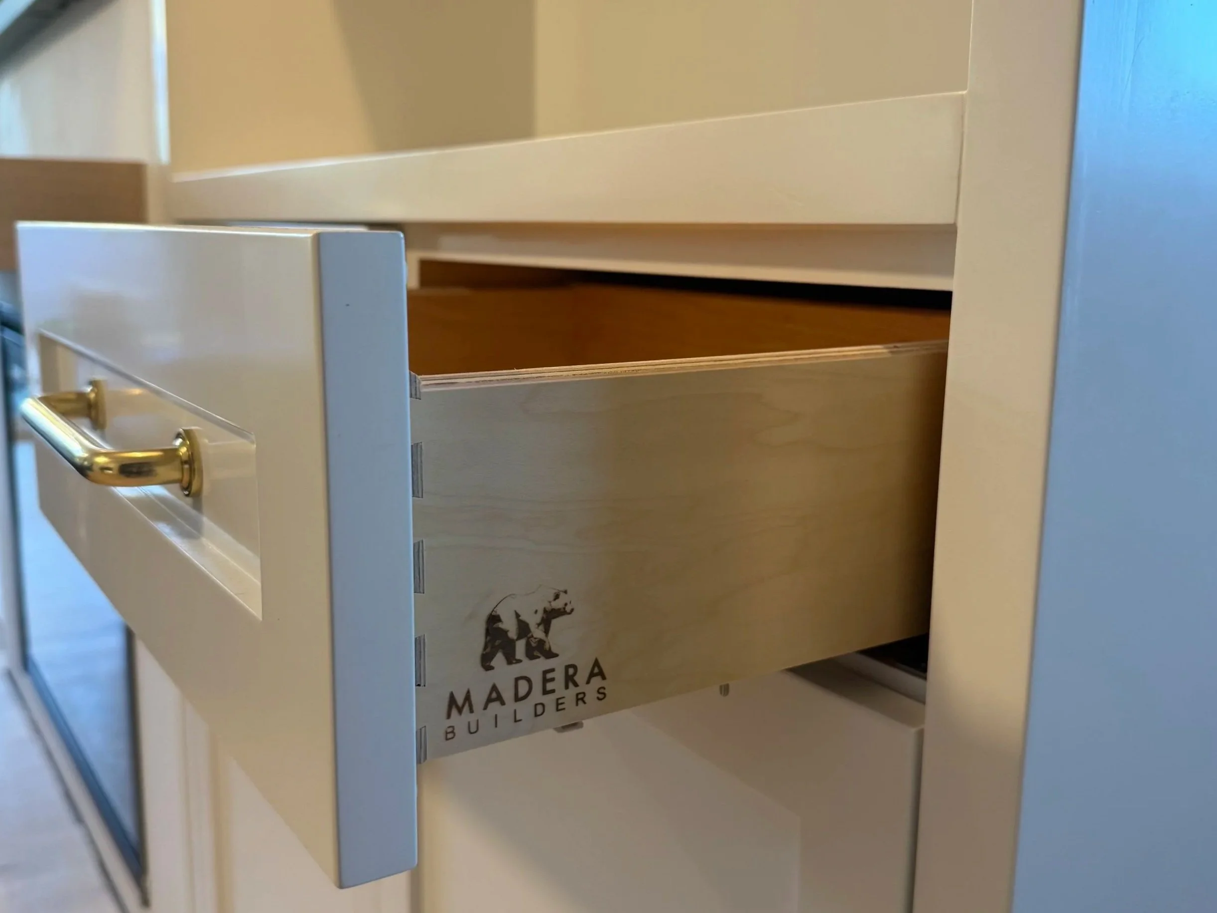 Close-up of a white wooden drawer with a gold handle, part of a furniture piece with a logo reading 'MADERA BUILDERS' on the side.