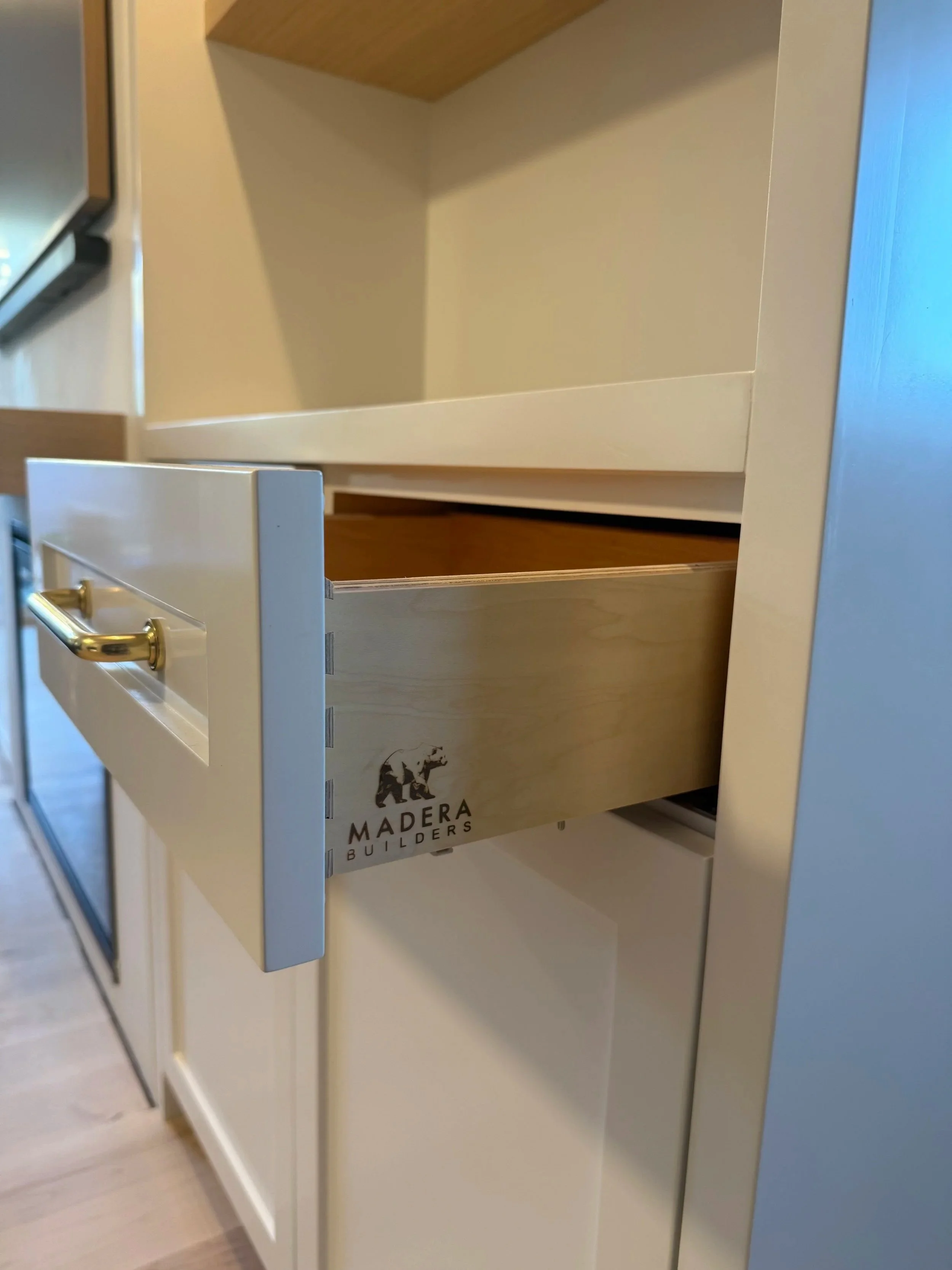 Close-up of an empty white cabinet with open drawers, showing a wooden interior and a logo reading 'MADERA BUILDERS' on the back panel.