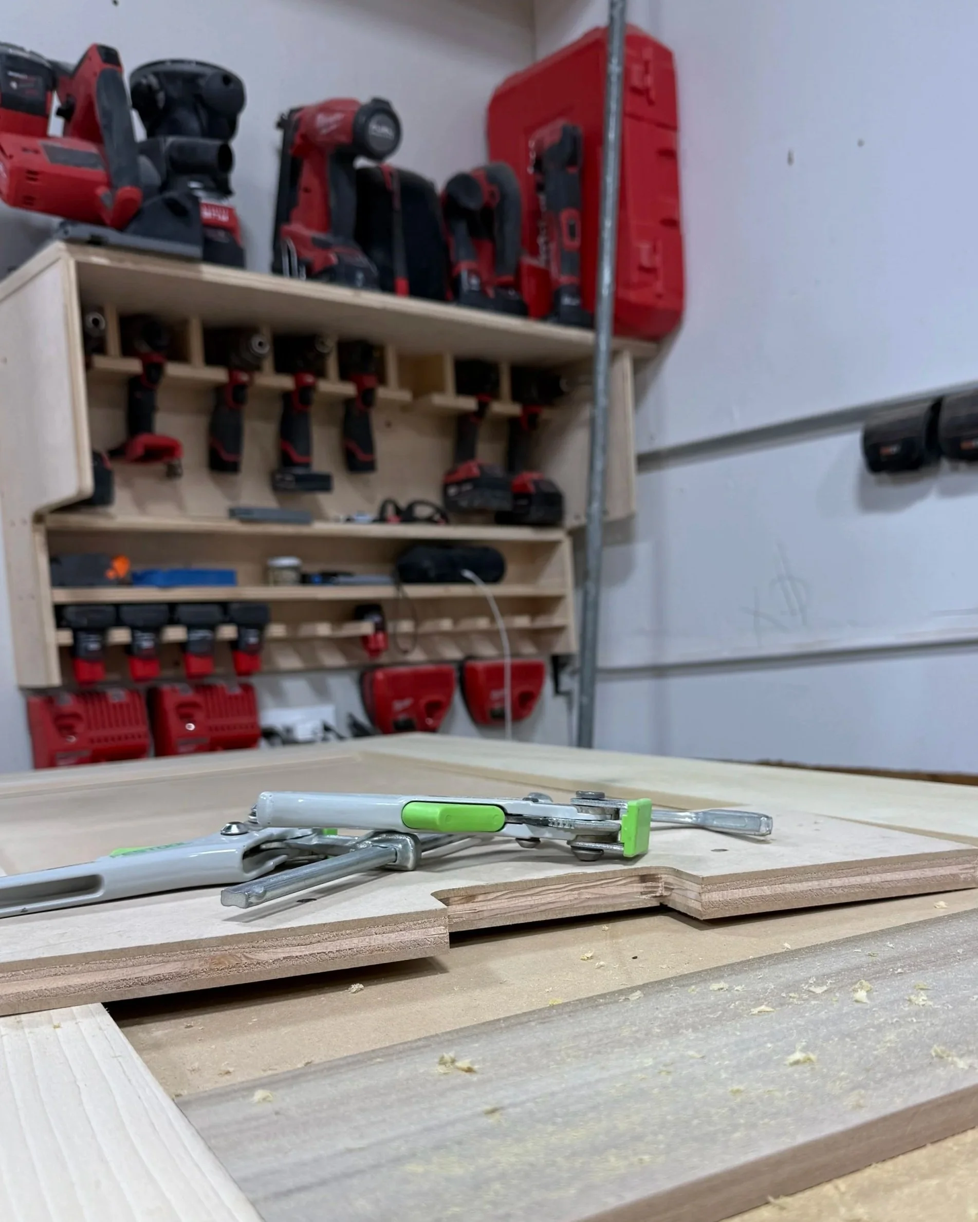 Worktable with clamps, along with stacked plywood sheets in a woodworking shop, tools and power tools on shelves in the background.