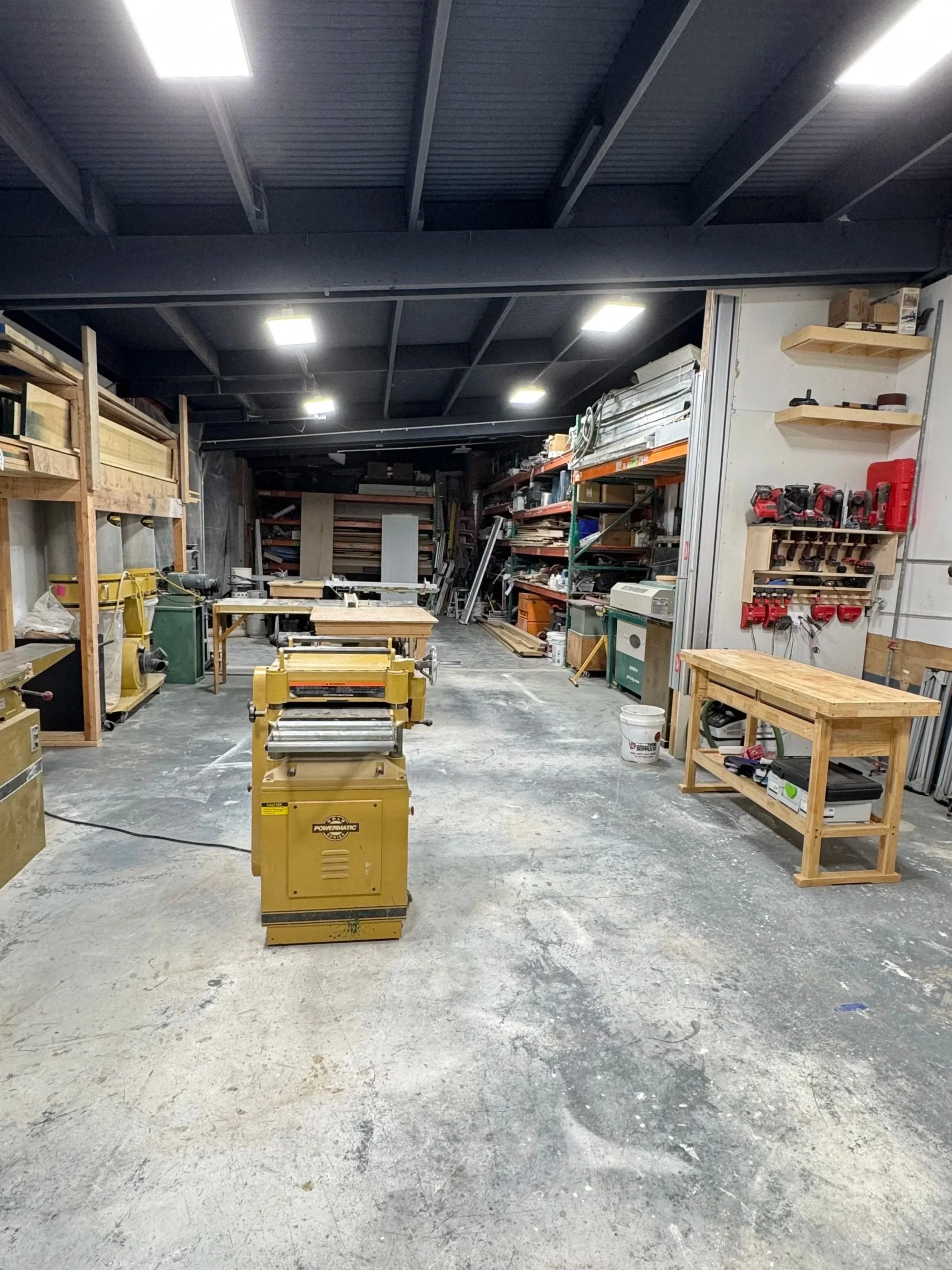 A workshop or storage room with shelves, woodworking tools, and equipment. The space has a concrete floor, black ceiling with fluorescent lights, and wooden workbenches and shelves.