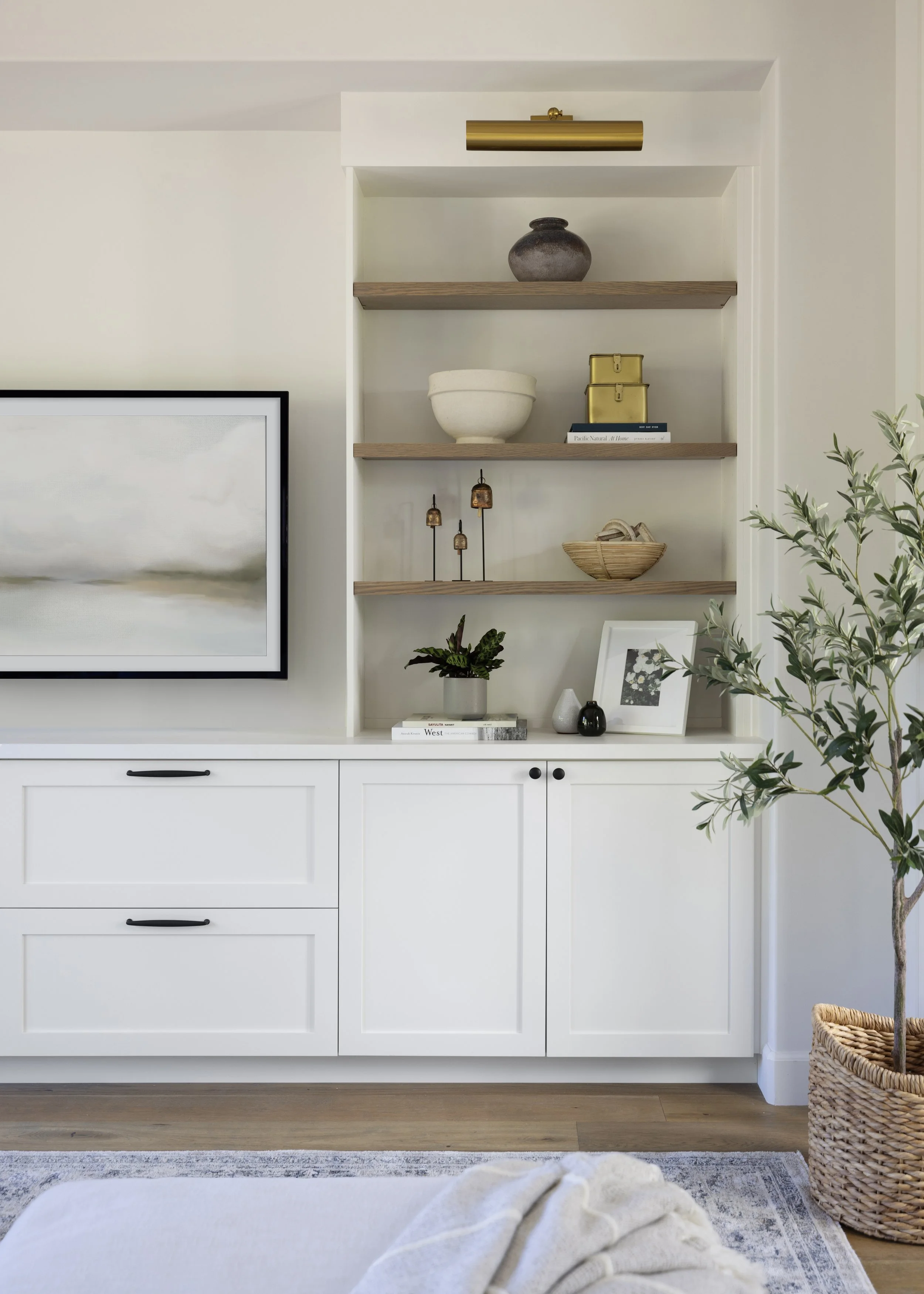 A modern interior with white cabinetry, decorative shelves holding vases and books, a large framed artwork, a potted plant, and a woven basket, with a bed and blanket in the foreground.