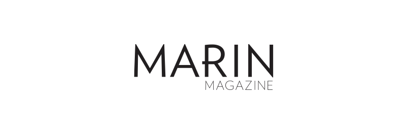 The cover of Marin Magazine featuring a charming outdoor scene of a woman in a plaid shirt and shorts holding a glass of wine, sitting on a stone wall by a body of water with trees and a house in the background.