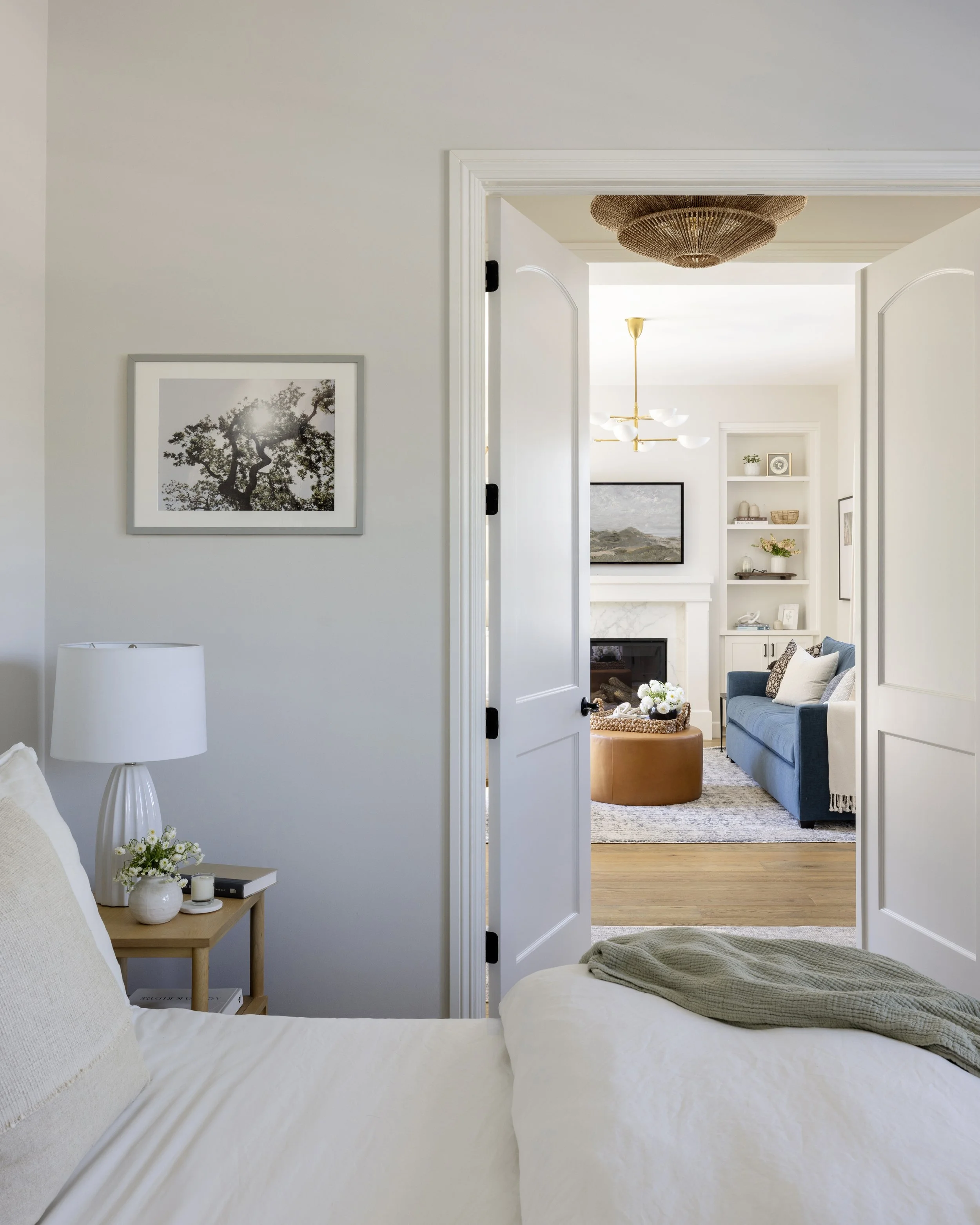 A view from a bedroom looking into a living room with open double doors. The bedroom has a beige bed with a white headboard, a small side table with a white lamp, and a framed black-and-white photo of a tree on the wall. The living room features a blue sofa with pillows, open shelving with decorative items, and a fireplace. The ceiling has a hanging light fixture. The overall decor is modern and minimalist with a neutral color palette.
