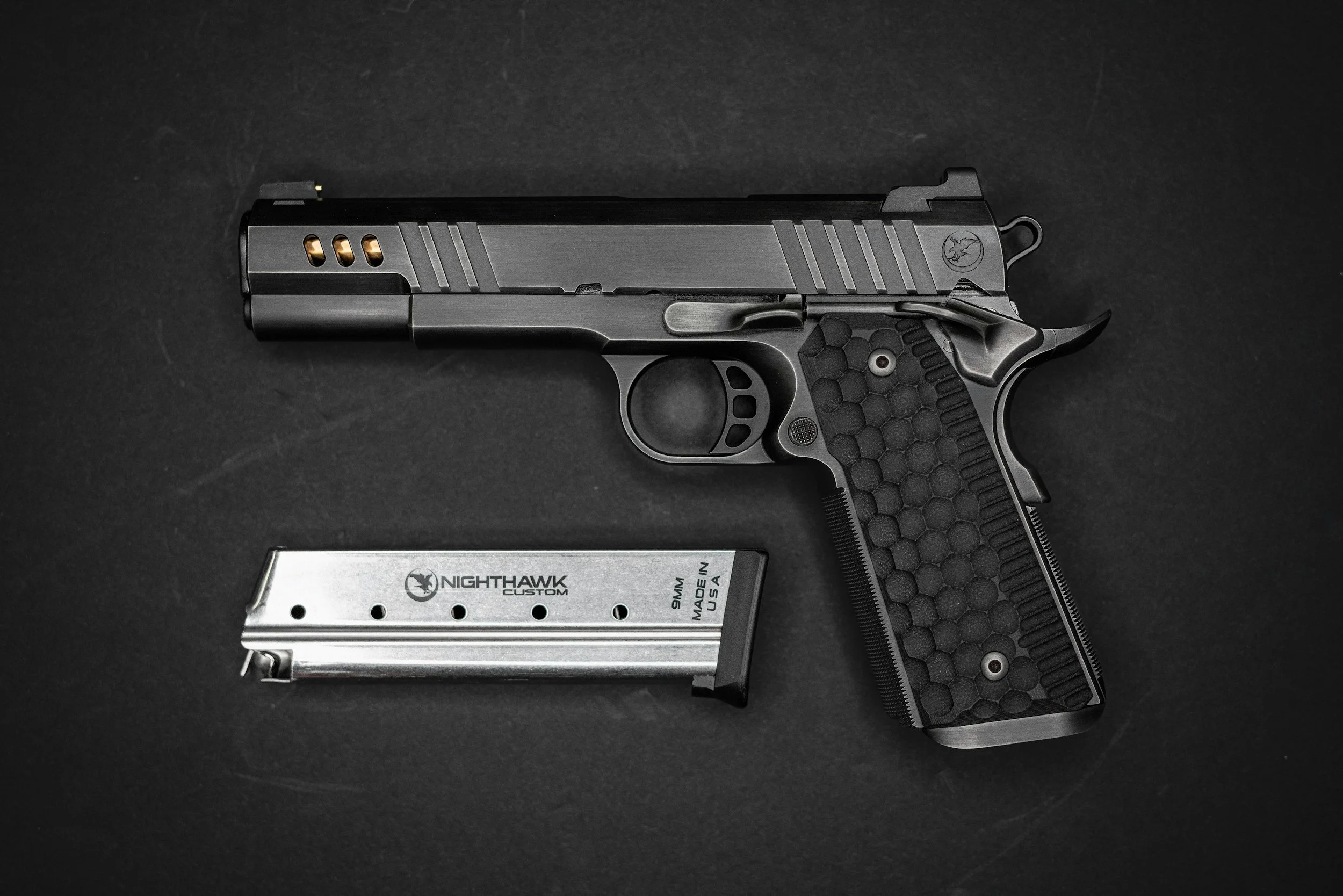 A black and gray semi-automatic handgun with a textured grip, placed on a dark surface alongside a silver magazine labeled "NightHawk Custom 9mm Made in USA."