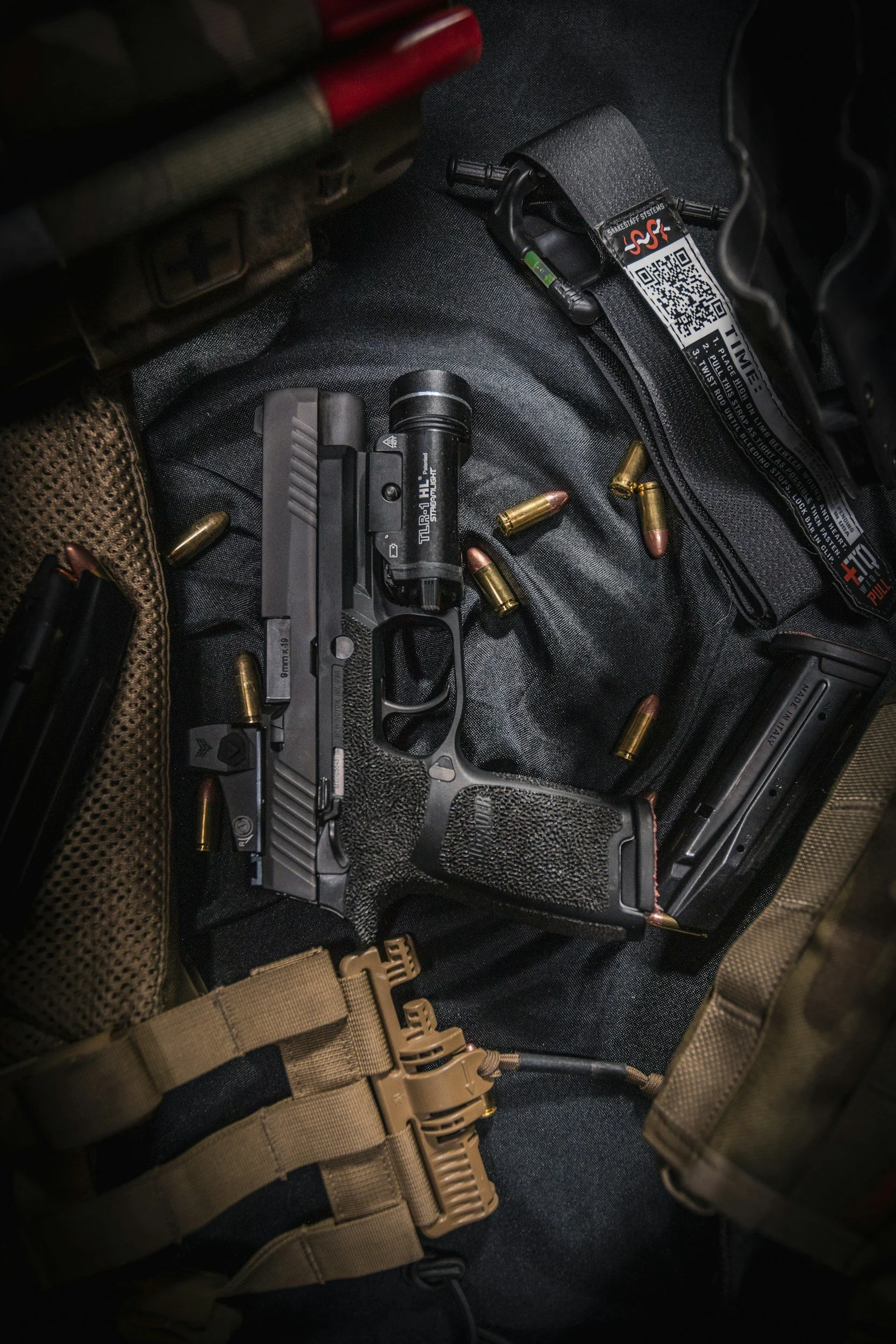 A firearm with several spent bullets around it, a military-style harness, and a laser sight attachment on a dark fabric surface.