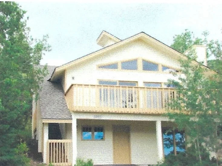 A two-story house with a balcony, beige siding, and a gable roof surrounded by trees.