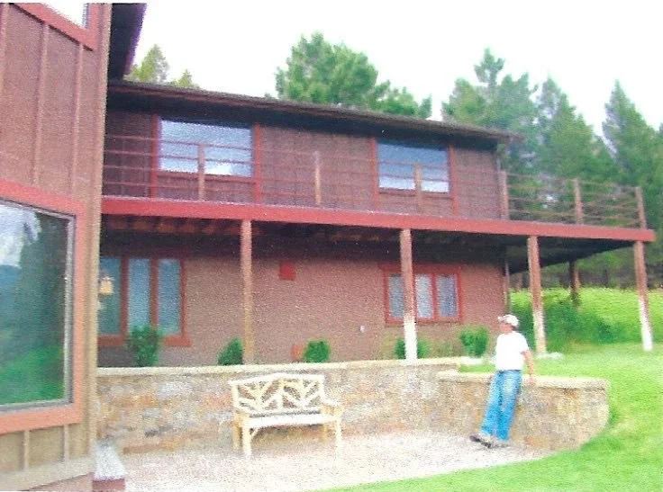 A person in a white hat and white shirt leaning against a stone wall outside a two-story house with a balcony, surrounded by green grass and trees.