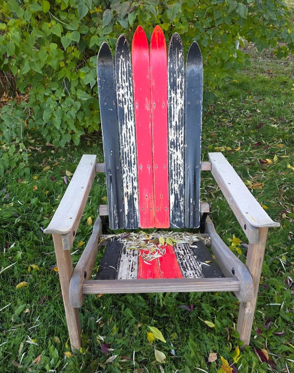 An Adirondack folding chair, using antique skis painted with a vintage red, white, and black color scheme, situated outdoors on grass.