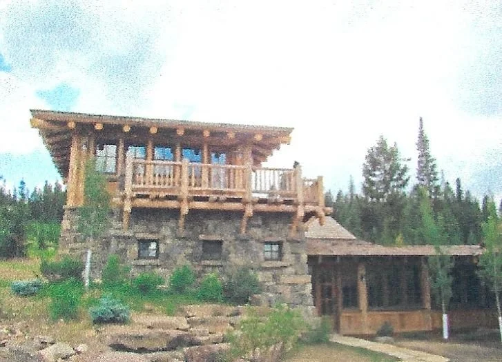 A rustic two-story cabin made of wood and stone surrounded by trees and greenery.
