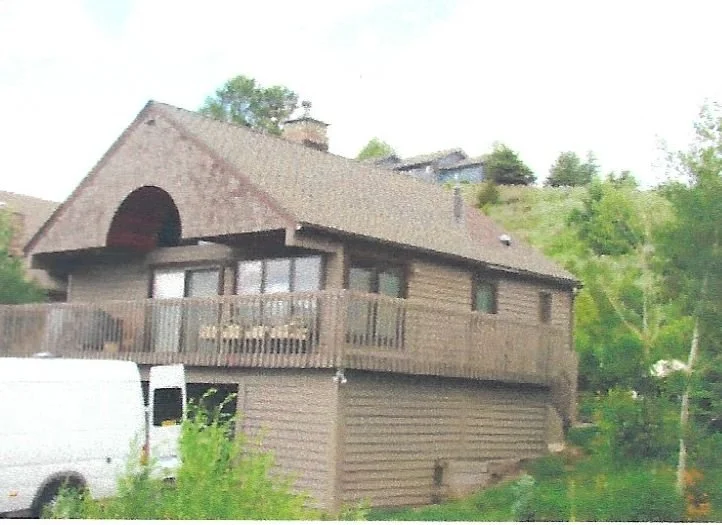 A brown house on stilts with a wooden deck, large windows, and a sloped roof, situated on a hillside with trees and a white van parked nearby.